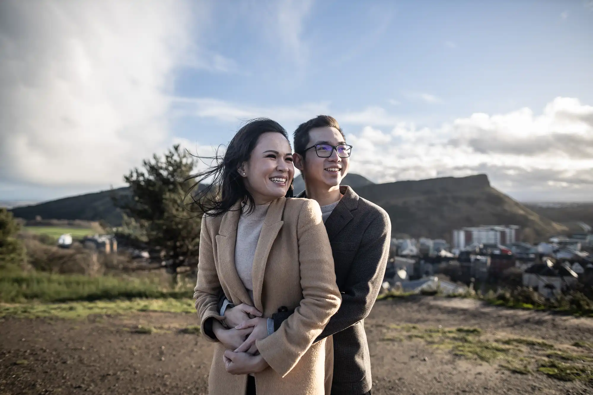 A couple stands embracing and smiling on a hilltop, with a view of a city and hilly landscape in the background under a partly cloudy sky.