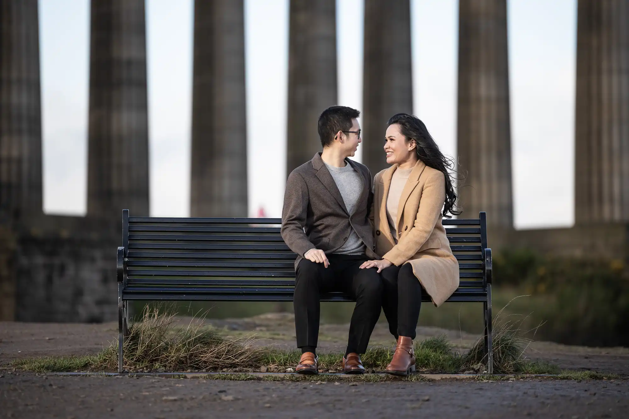 A man and woman sit on a black bench, outdoors, in front of large pillars. They are facing each other, smiling, with the woman resting her hand on the man’s knee.