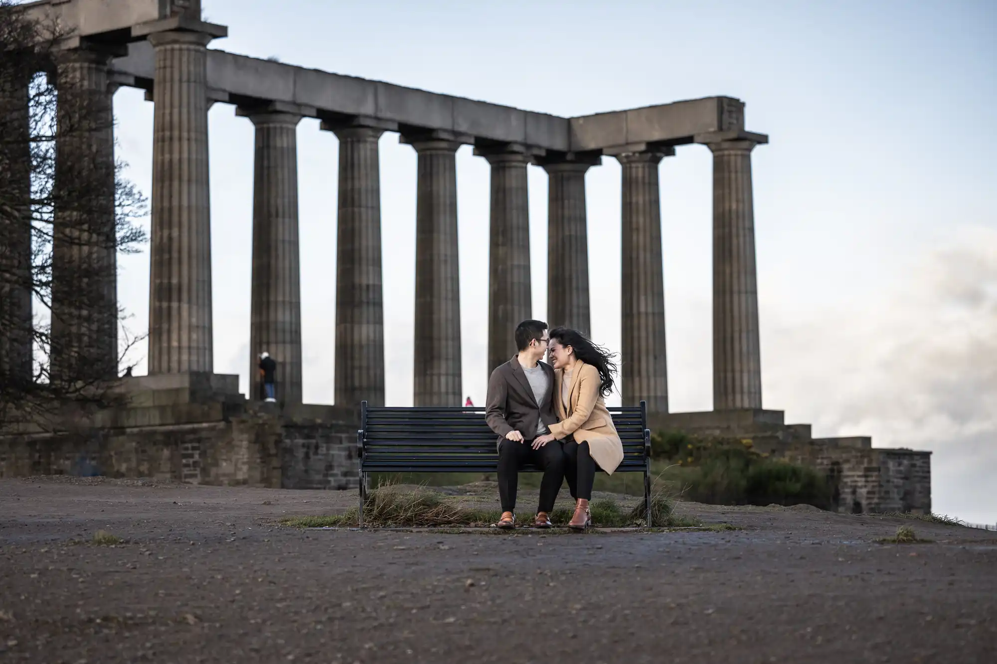 A couple sits closely on a bench in front of a historic columned structure, with one person whispering into the other's ear.