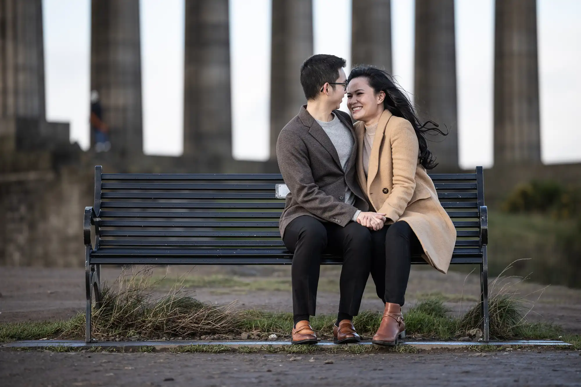 A couple sits on a black bench, smiling and holding hands. They are outdoors in front of historical stone columns on a cloudy day.