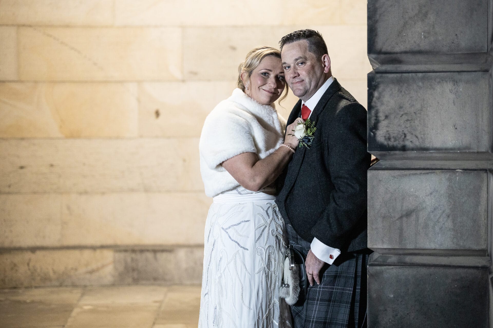 A couple dressed in formal attire poses together, with the woman in a white dress and shawl and the man in a dark jacket and plaid kilt, standing against a stone wall.