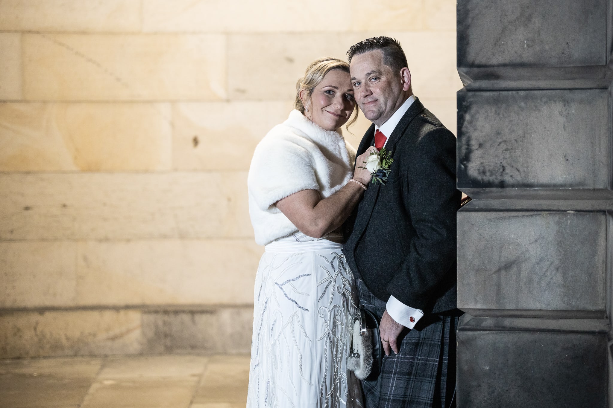 A couple dressed in formal attire poses together, with the woman in a white dress and shawl and the man in a dark jacket and plaid kilt, standing against a stone wall.