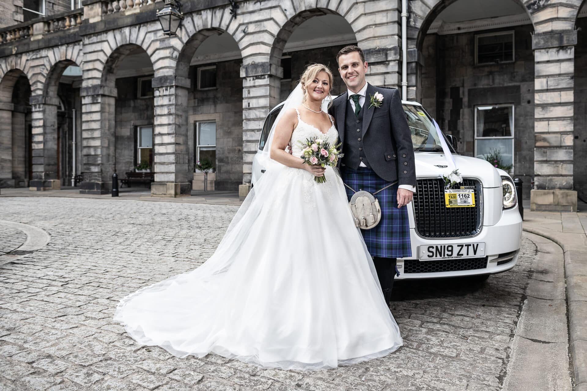 A bride and groom in wedding attire stand in front of a white car with a visible license plate, outside a stone building with arches.