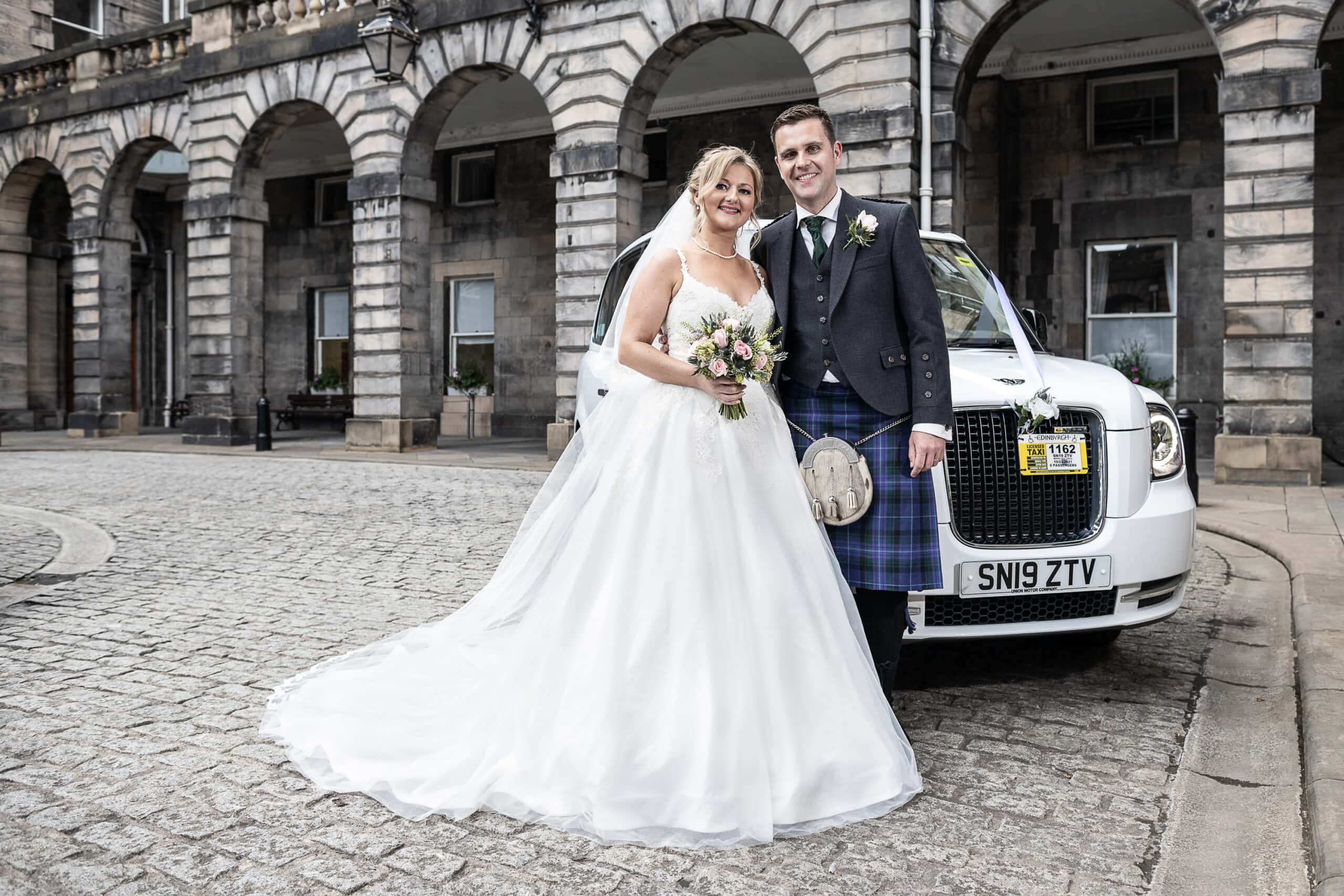 A bride and groom in wedding attire stand in front of a white car with a visible license plate, outside a stone building with arches.