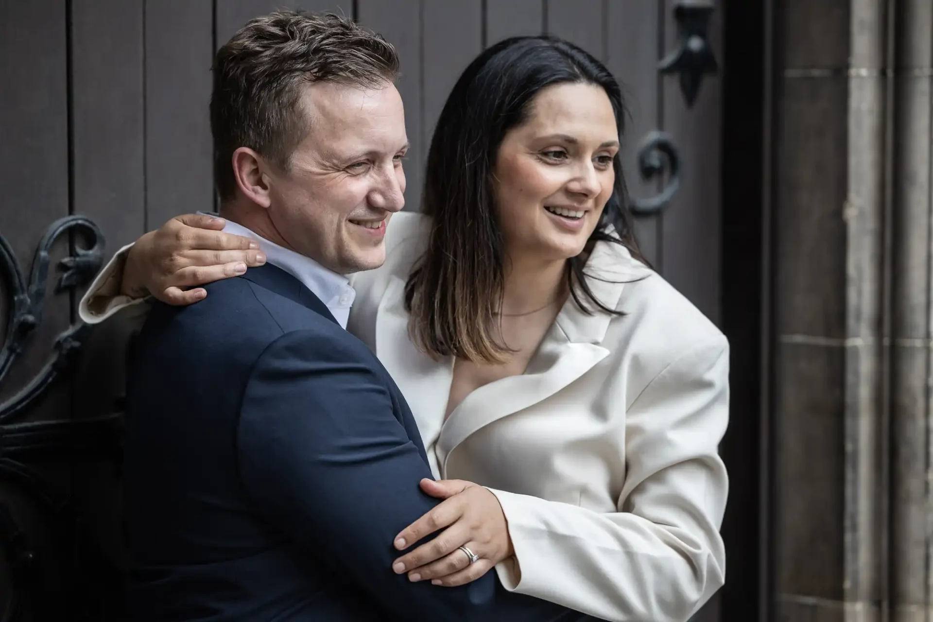 A man in a dark suit and a woman in a white outfit stand closely together, smiling and facing slightly to the right. She has her arm around his shoulder.
