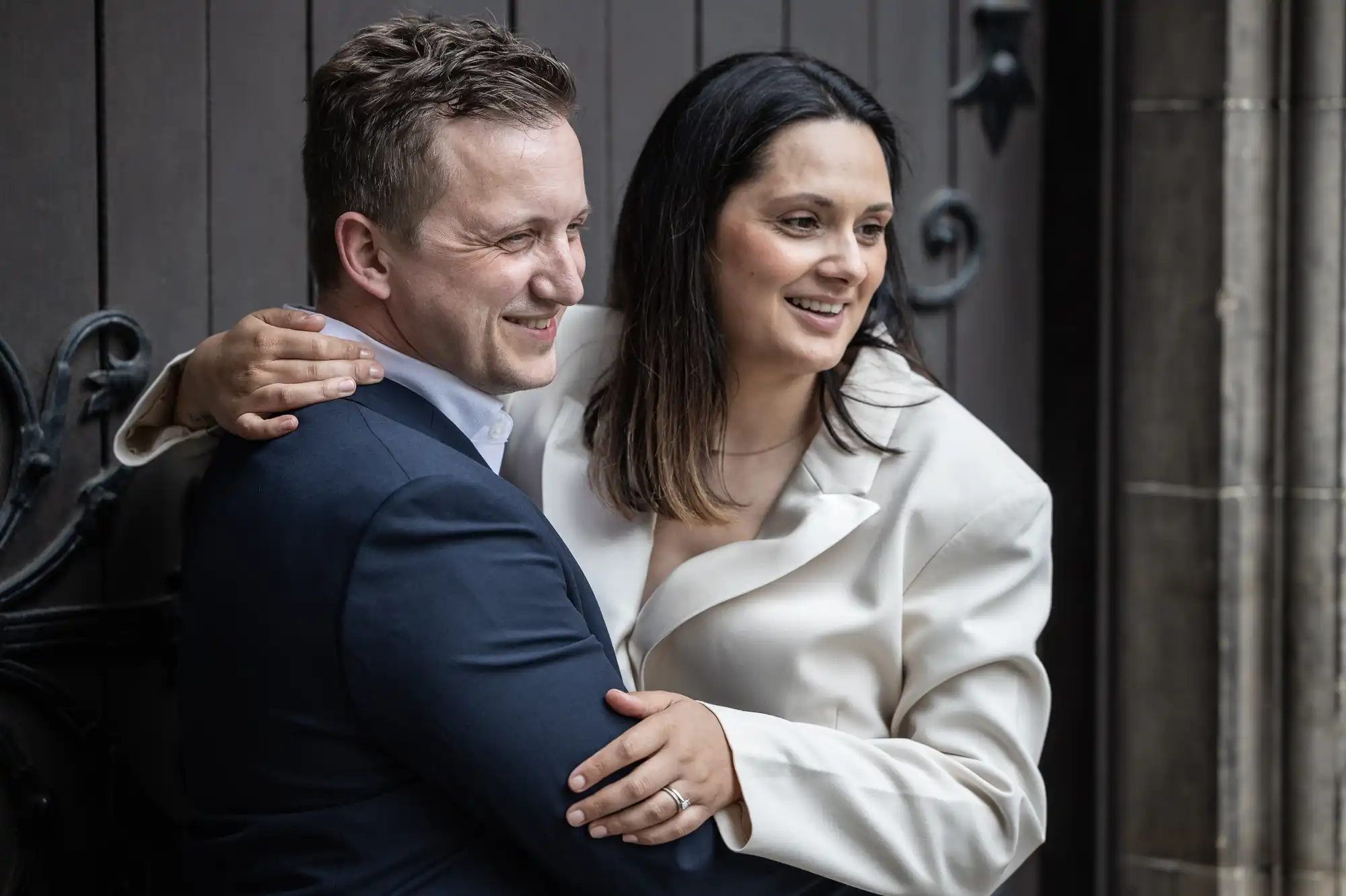 A man in a dark suit and a woman in a white outfit stand closely together, smiling and facing slightly to the right. She has her arm around his shoulder.