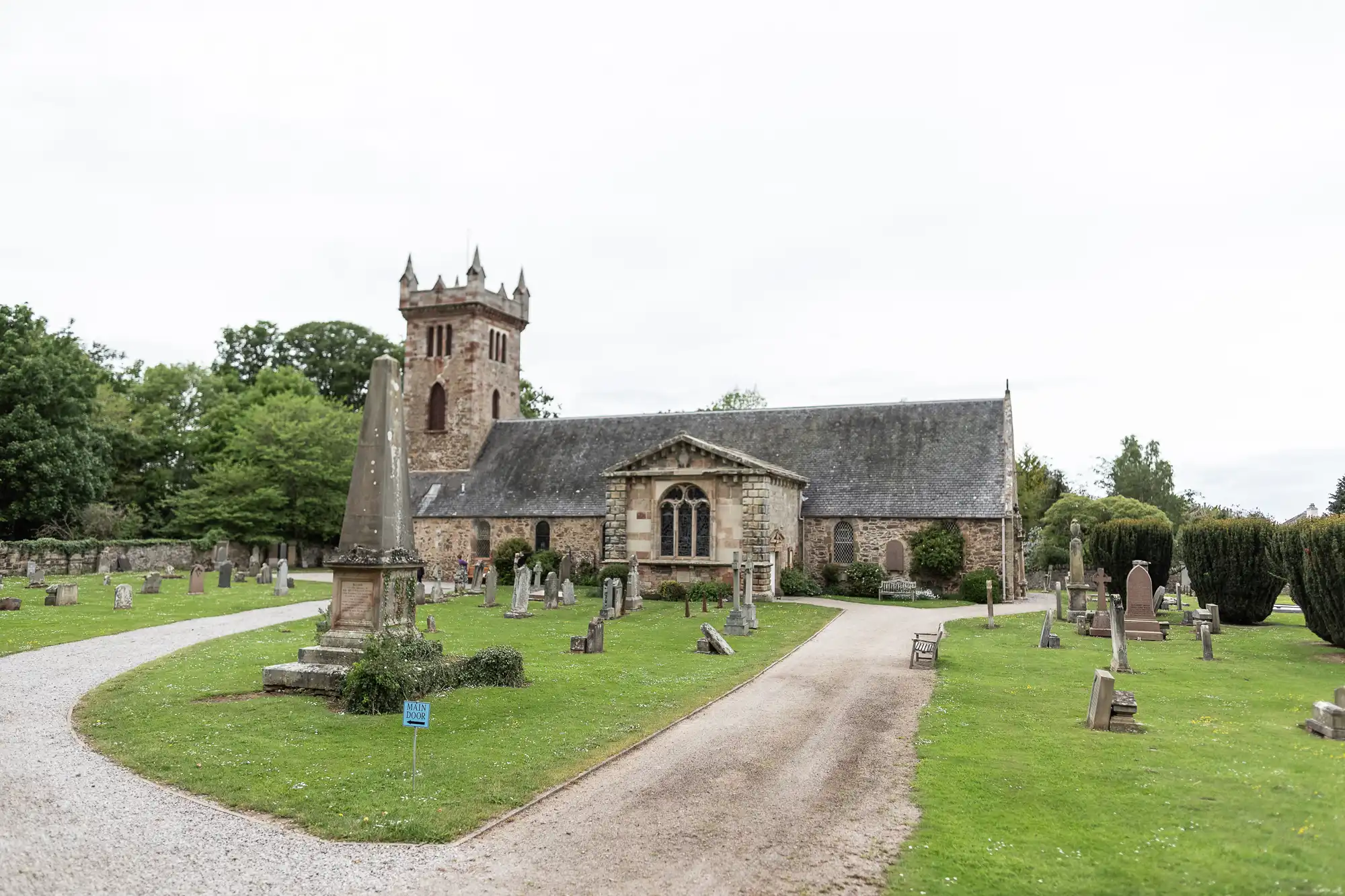 A historic stone church with a tall steeple is surrounded by a grassy cemetery with various gravestones and a pathway leading up to the entrance.