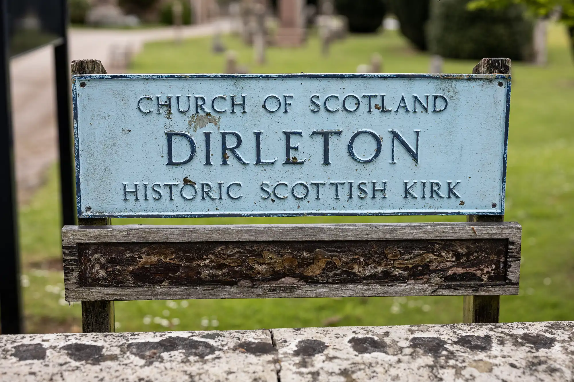 Sign reading "Church of Scotland Dirleton Historic Scottish Kirk" with a blurred background of grass and stone structures.