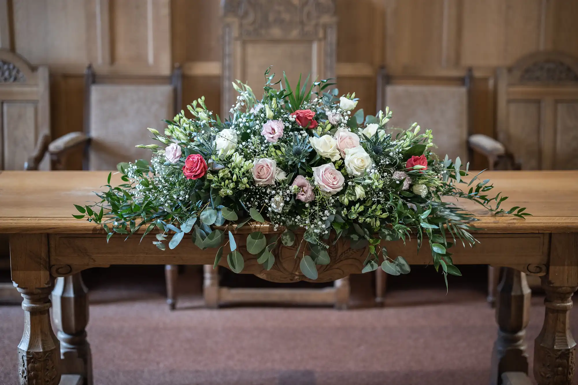 Floral arrangement with roses and greenery on a wooden table in a room with wooden panels and upholstered chairs.