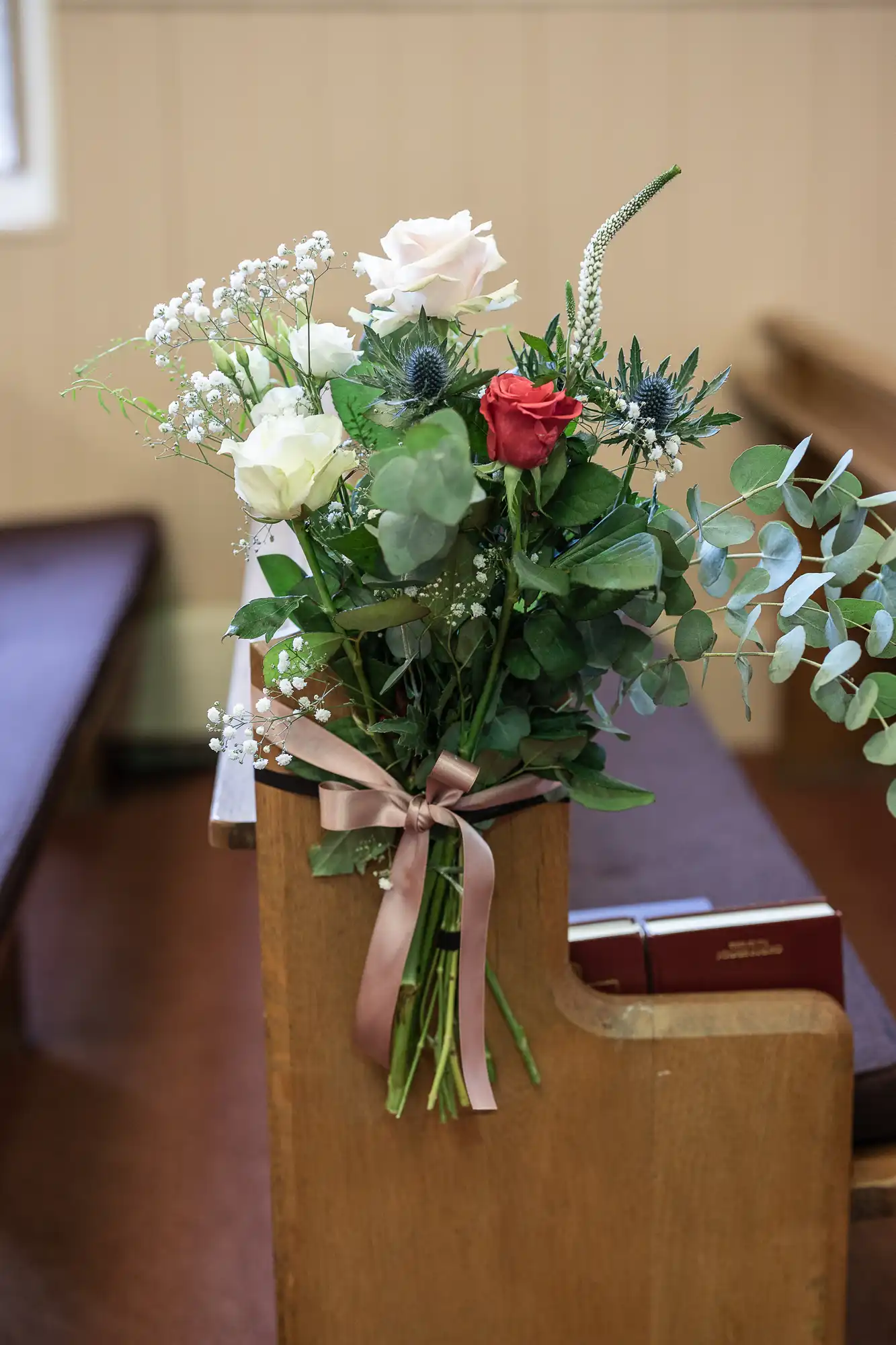 A bouquet of white, pink, and red flowers tied with a pink ribbon is attached to an end pew in a church, with two hymn books placed on the seat beside it.