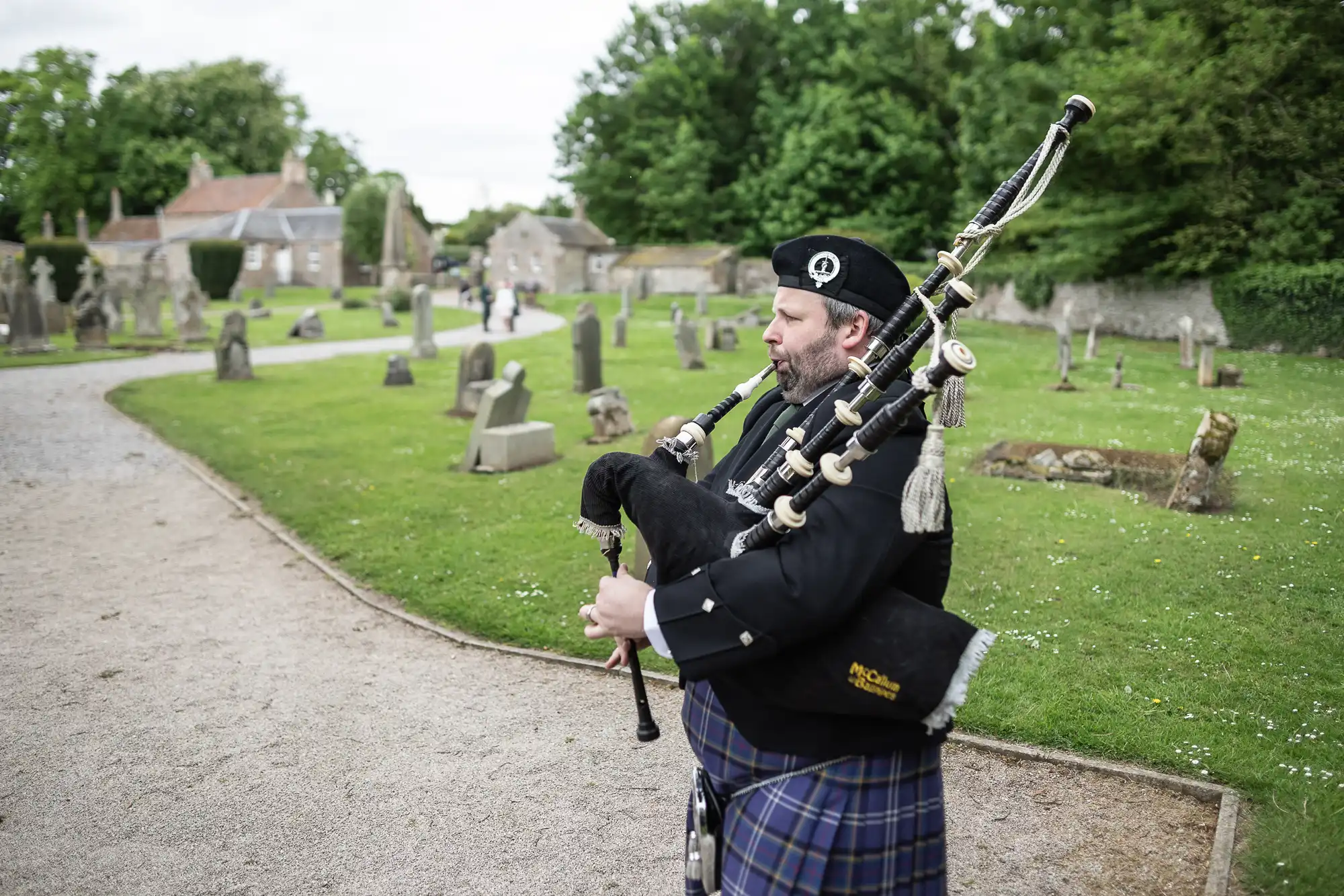 A man wearing traditional Scottish attire plays the bagpipes in an outdoor setting with grass, trees, and tombstones in the background.