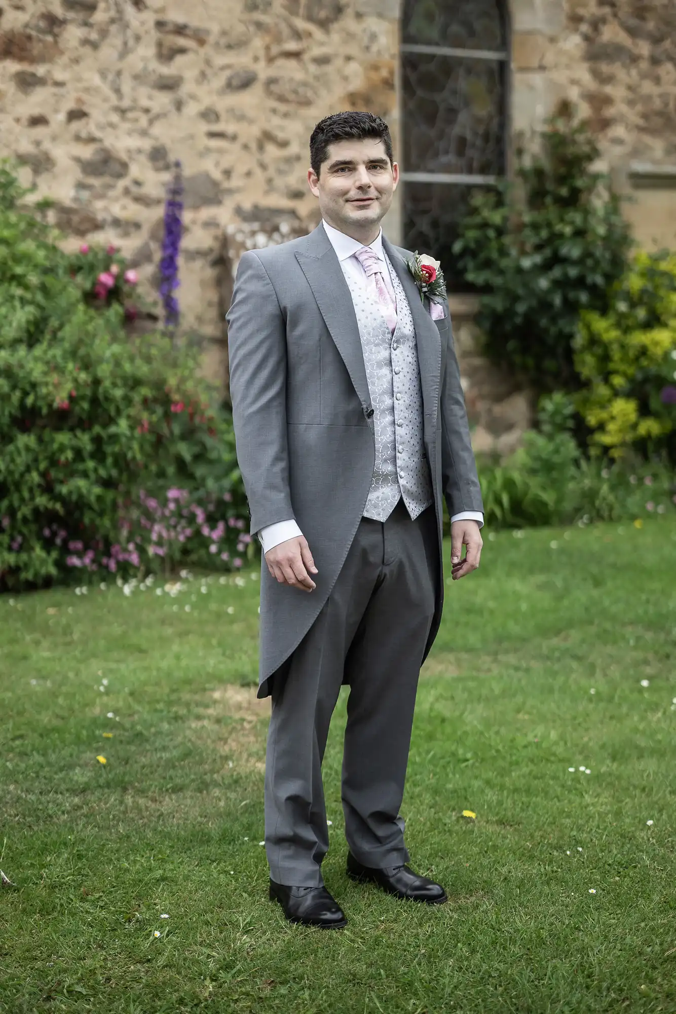 A man in a gray suit with a light pink tie and floral boutonniere stands on a lawn in front of a stone building with green foliage and flowers in the background.