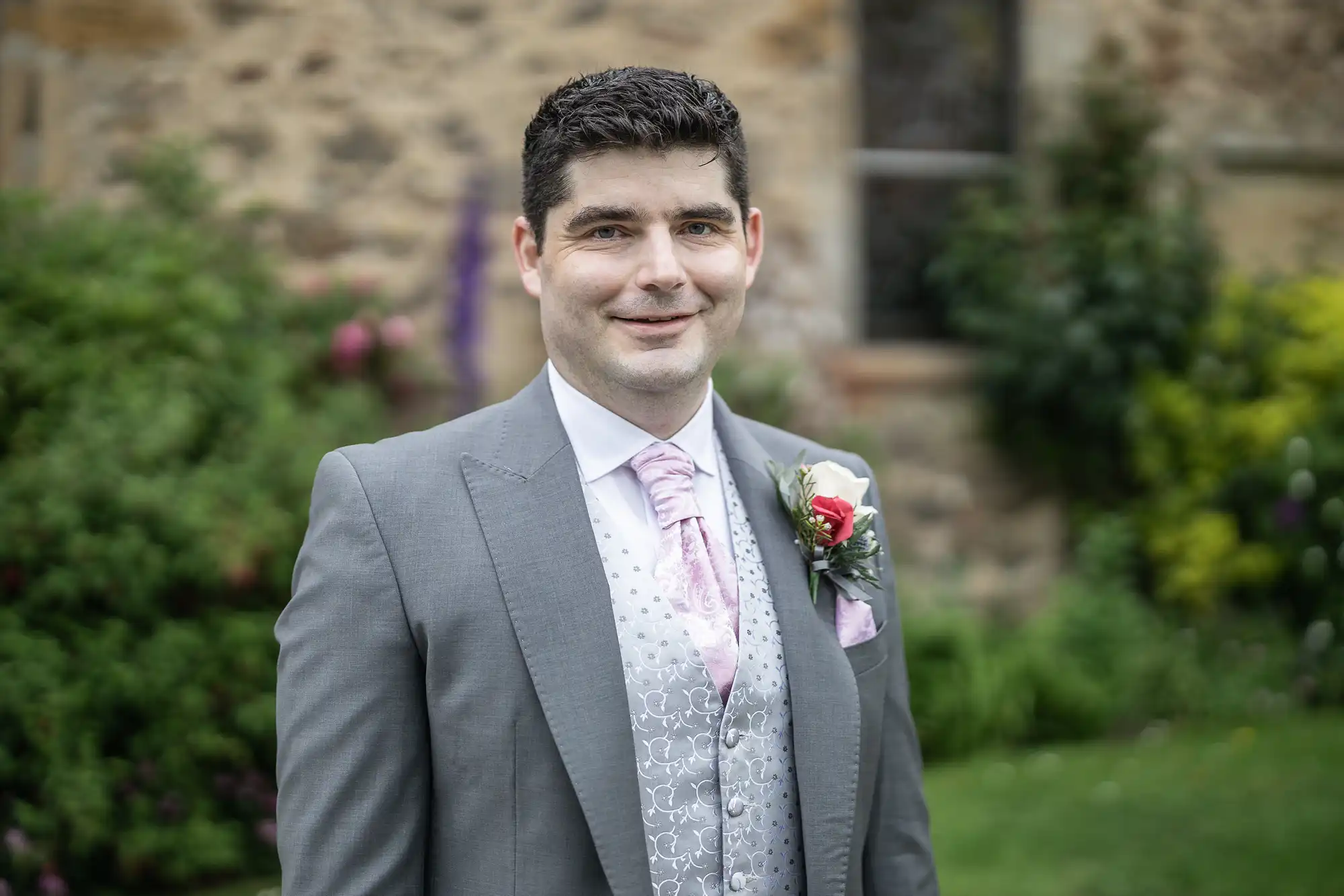 A man wearing a gray suit with a light pink tie and a floral boutonniere stands outdoors in front of a stone building and greenery.