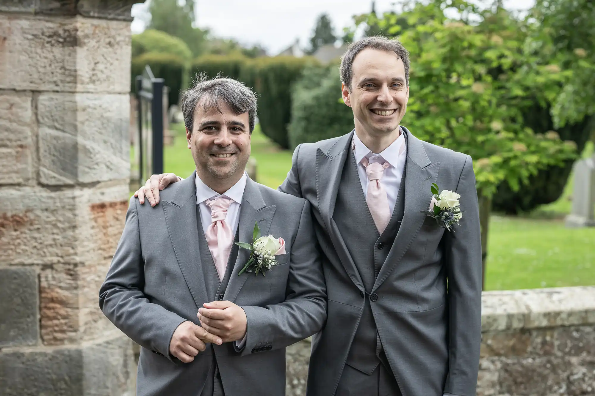 Two men in matching grey suits with pink ties and rose boutonnieres smile while standing outdoors by a stone building. One man has his arm around the other. Trees and greenery are visible in the background.