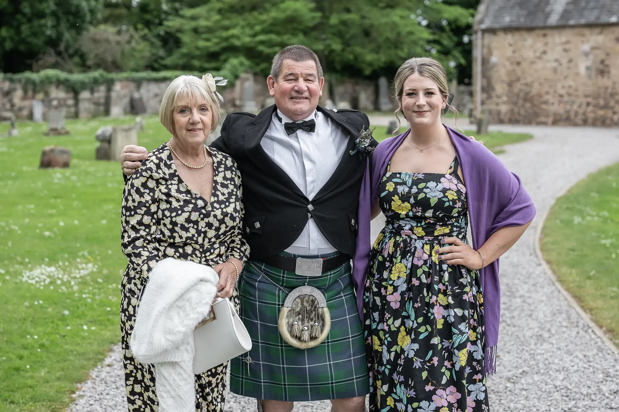 Three people are posing together: a woman in a patterned dress holding a white purse, a man in a kilt and bow tie, and a woman in a floral dress with a purple shawl, standing on a gravel path.