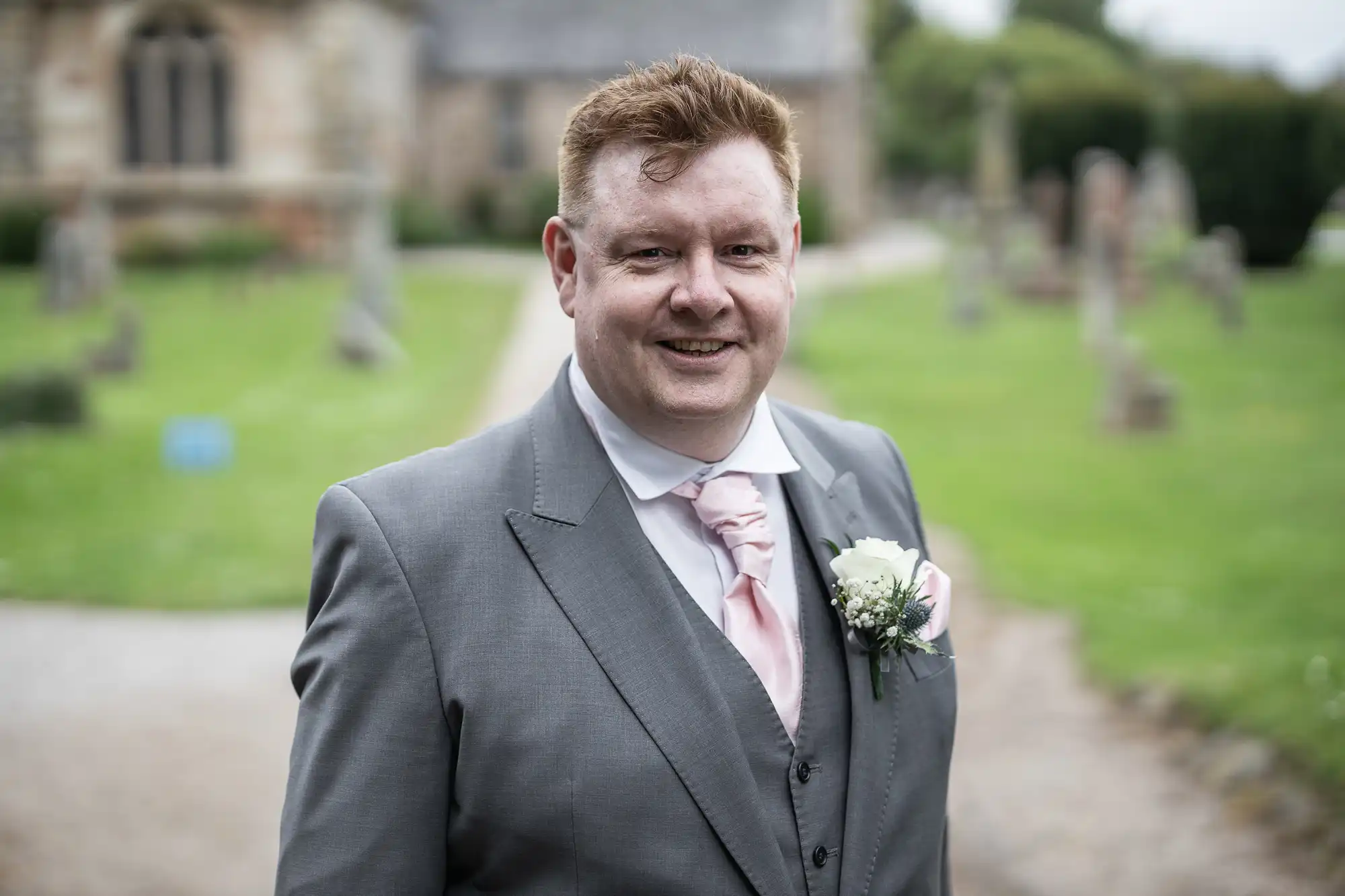 A man in a grey suit with a pink tie and a white floral boutonniere stands outdoors, smiling at the camera. Blurred background of a church and greenery.