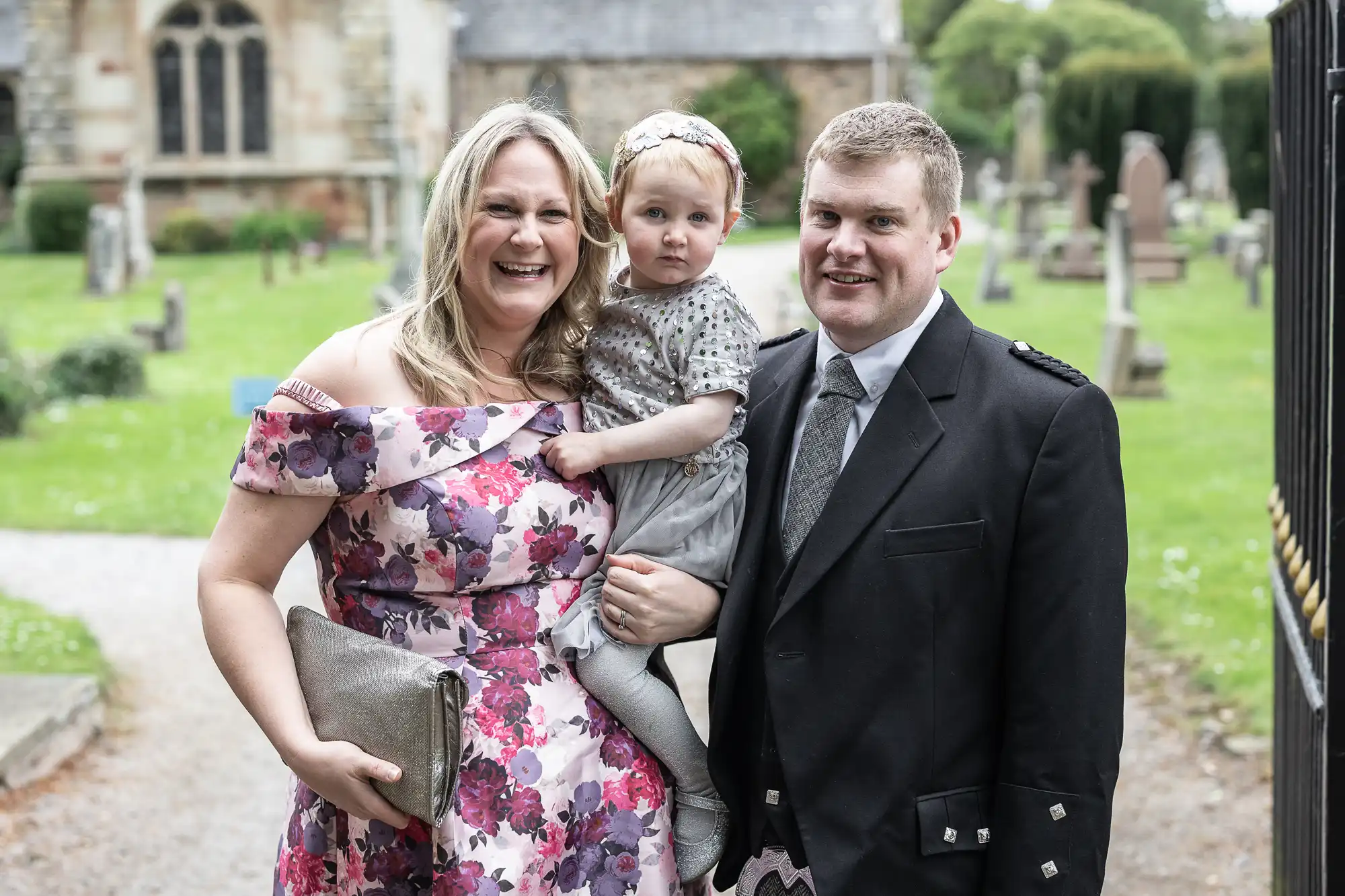 A woman in a floral dress, a man in a black suit, and a toddler in a gray dress pose together outdoors in front of a church and gravestones.