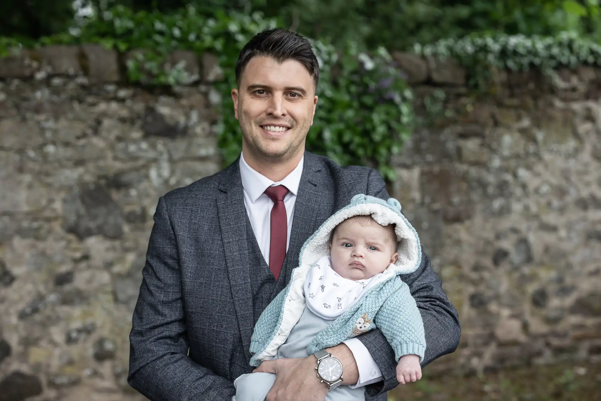 A man in a suit holds a baby dressed in a light blue sweater with bear ears on the hood, standing in front of a stone wall with greenery.