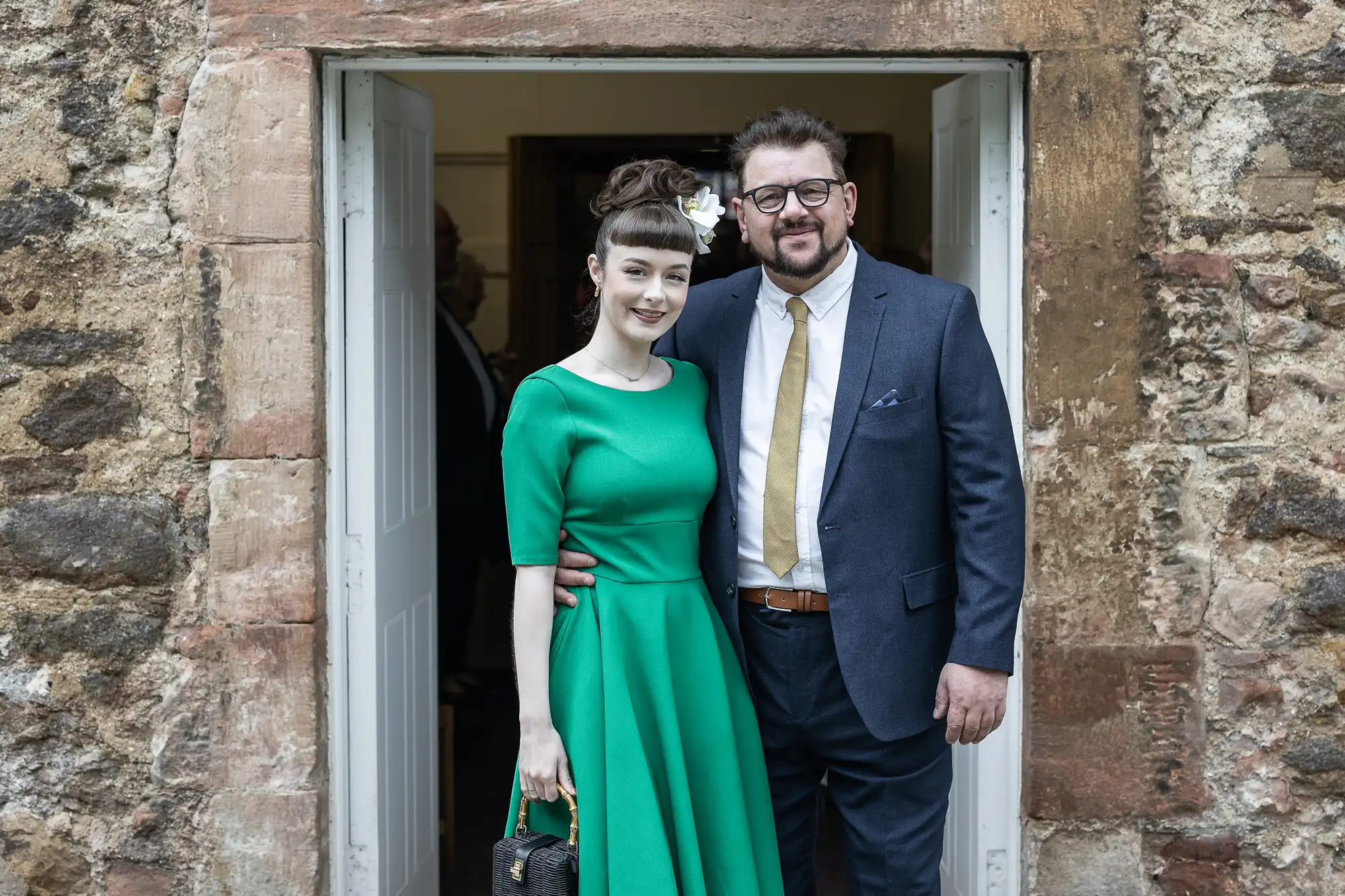 A smiling couple stands in a doorway; the woman wears a green dress with a floral hair accessory, and the man wears glasses, a dark suit, and a yellow tie. They appear to be at an event.