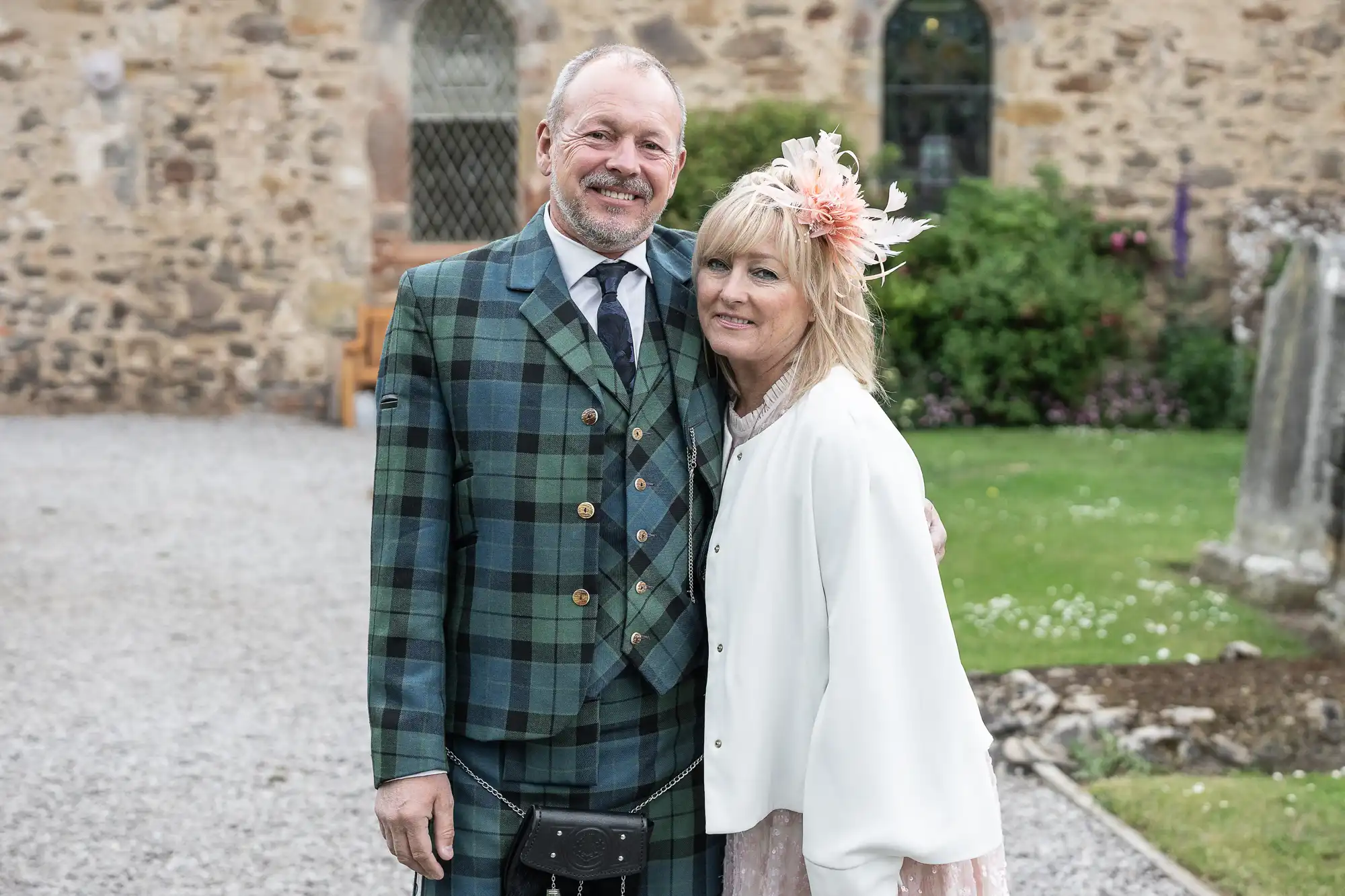 A couple stands together outdoors in front of a stone building. The man is wearing a green plaid kilt outfit, and the woman is wearing a white jacket with a floral hair accessory.