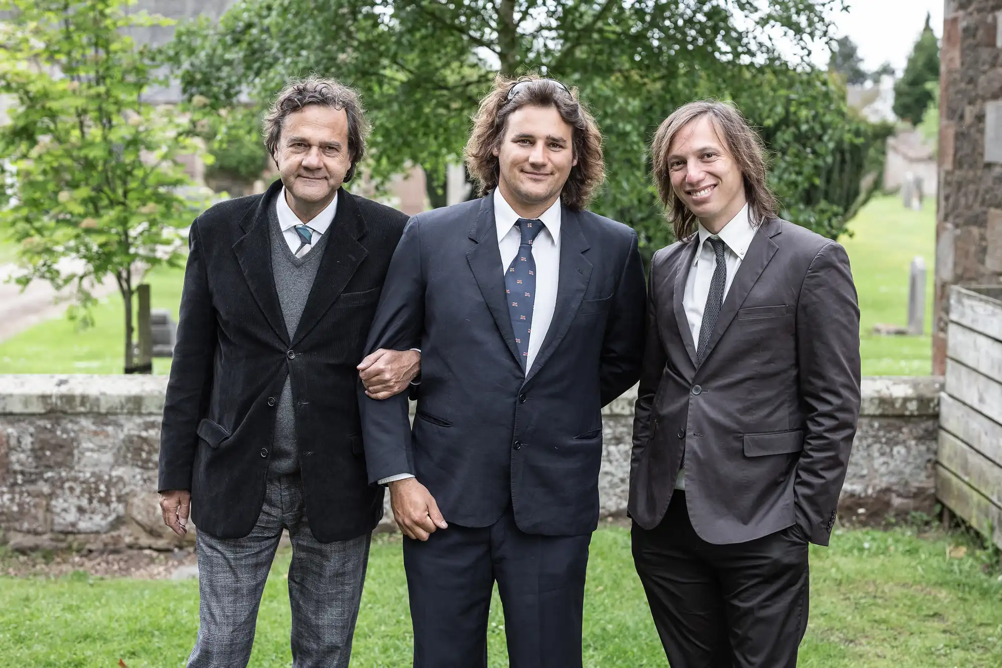 Three men, dressed in suits, stand together outdoors on a grassy area with stone buildings and trees in the background. The man in the center is linking arms with the other two men.