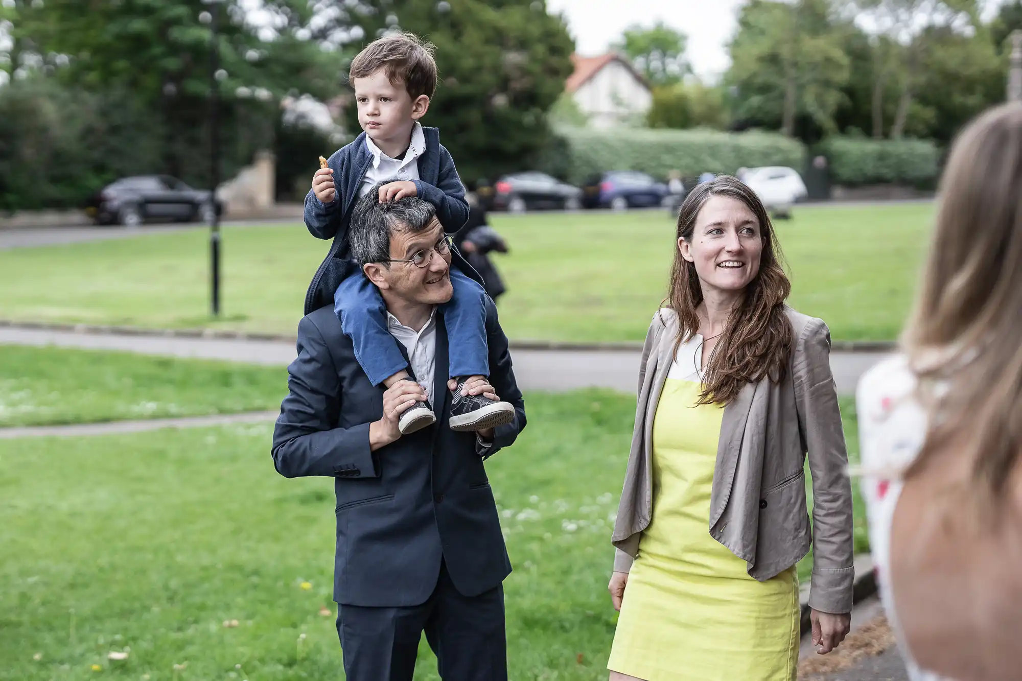 A man carries a young boy on his shoulders, holding his legs while the boy eats a snack. A woman in a yellow dress stands next to them, all outdoors in a park setting.