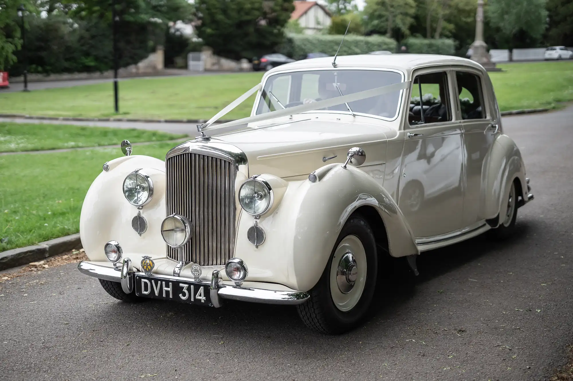 A vintage white car, likely from the mid-20th century, parked on a paved roadway with greenery and trees in the background. License plate reads "DVH 314.