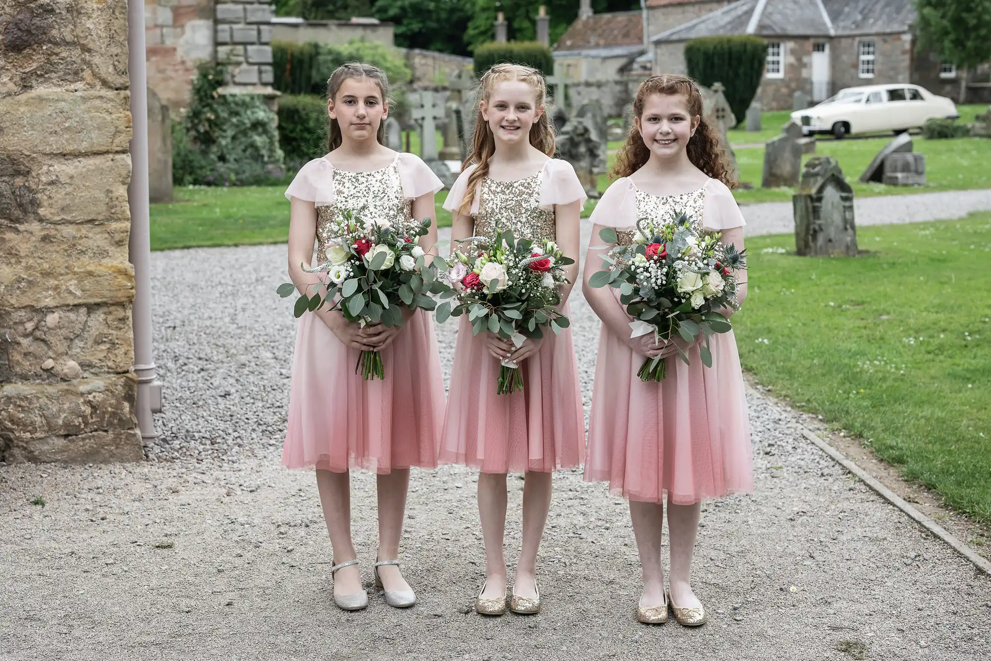 Three girls in matching pink dresses holding flower bouquets stand in a garden with a stone building and an old car in the background.