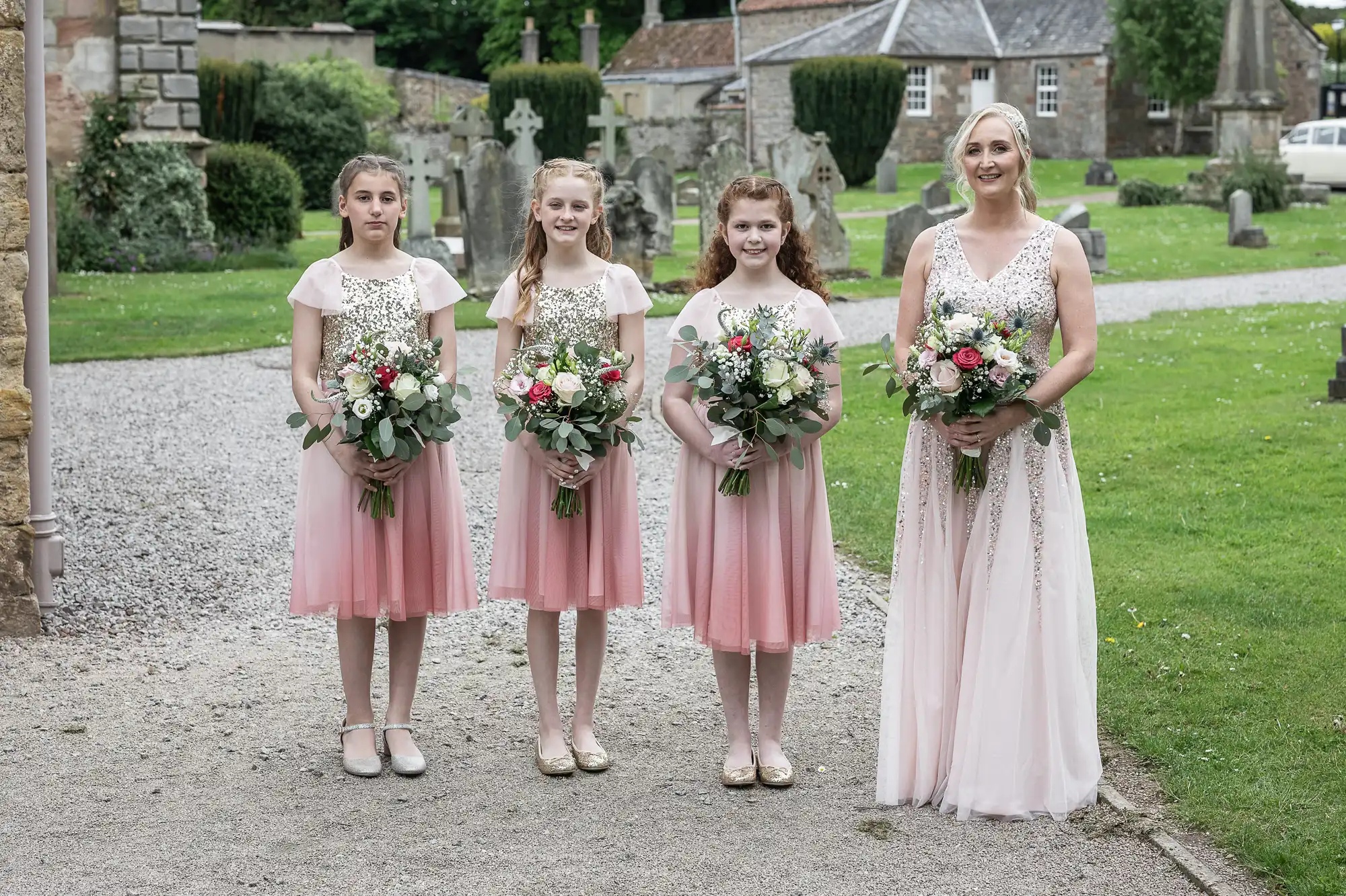 A bride in a white dress stands with three bridesmaids in pink dresses, each holding a bouquet, posing outdoors in front of a pathway and greenery.