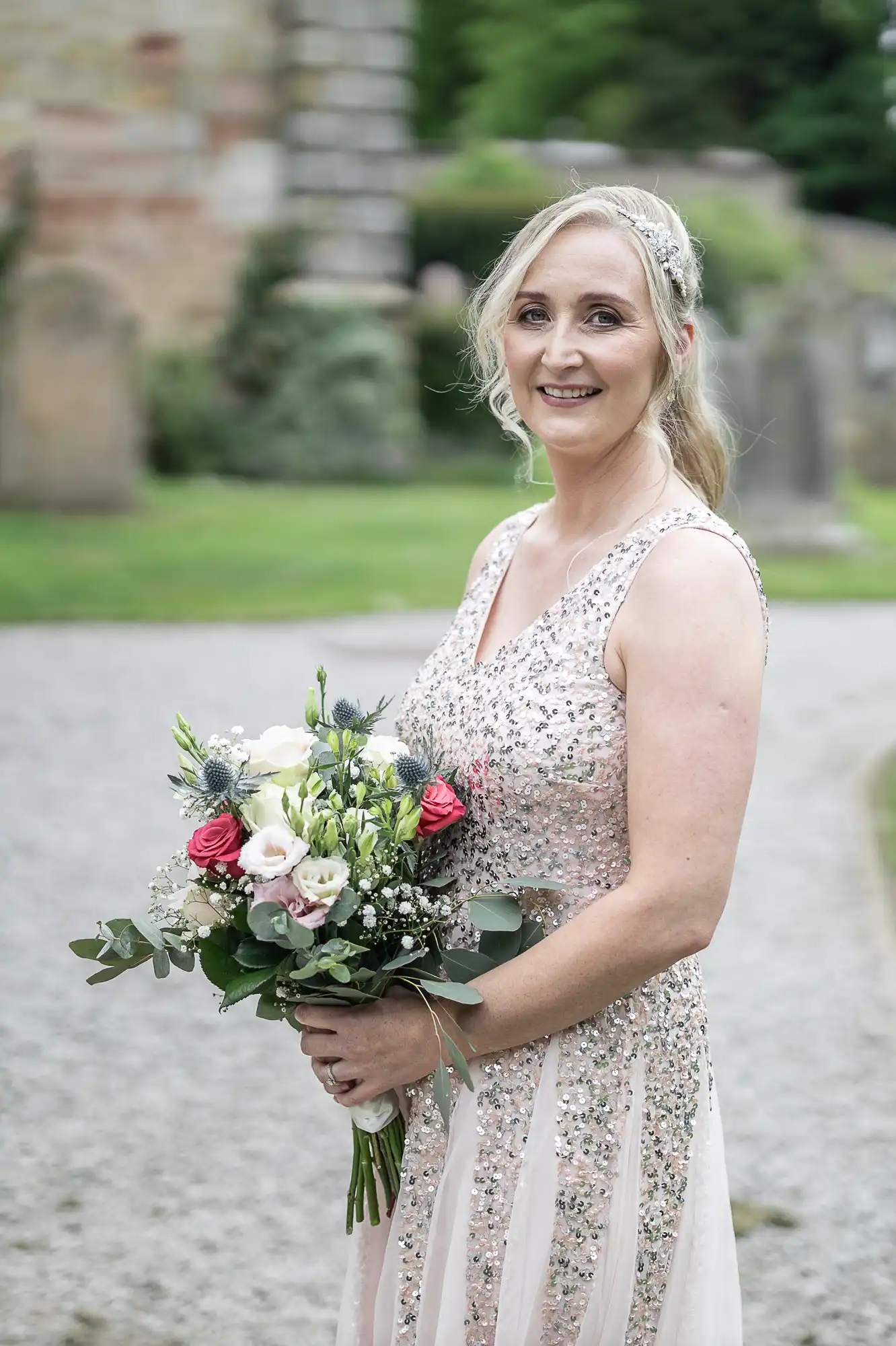 A woman in a sparkly, sleeveless dress holds a bouquet of flowers and smiles at the camera while standing outside on a gravel path.