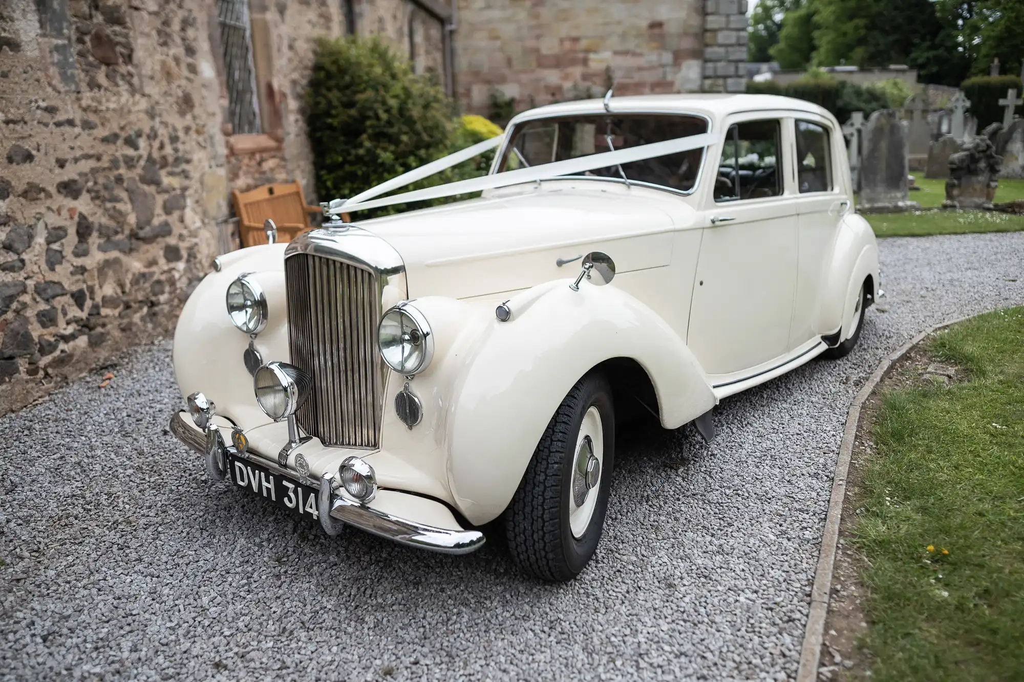 A white vintage car decorated with a ribbon is parked on a gravel path next to a stone building and greenery.