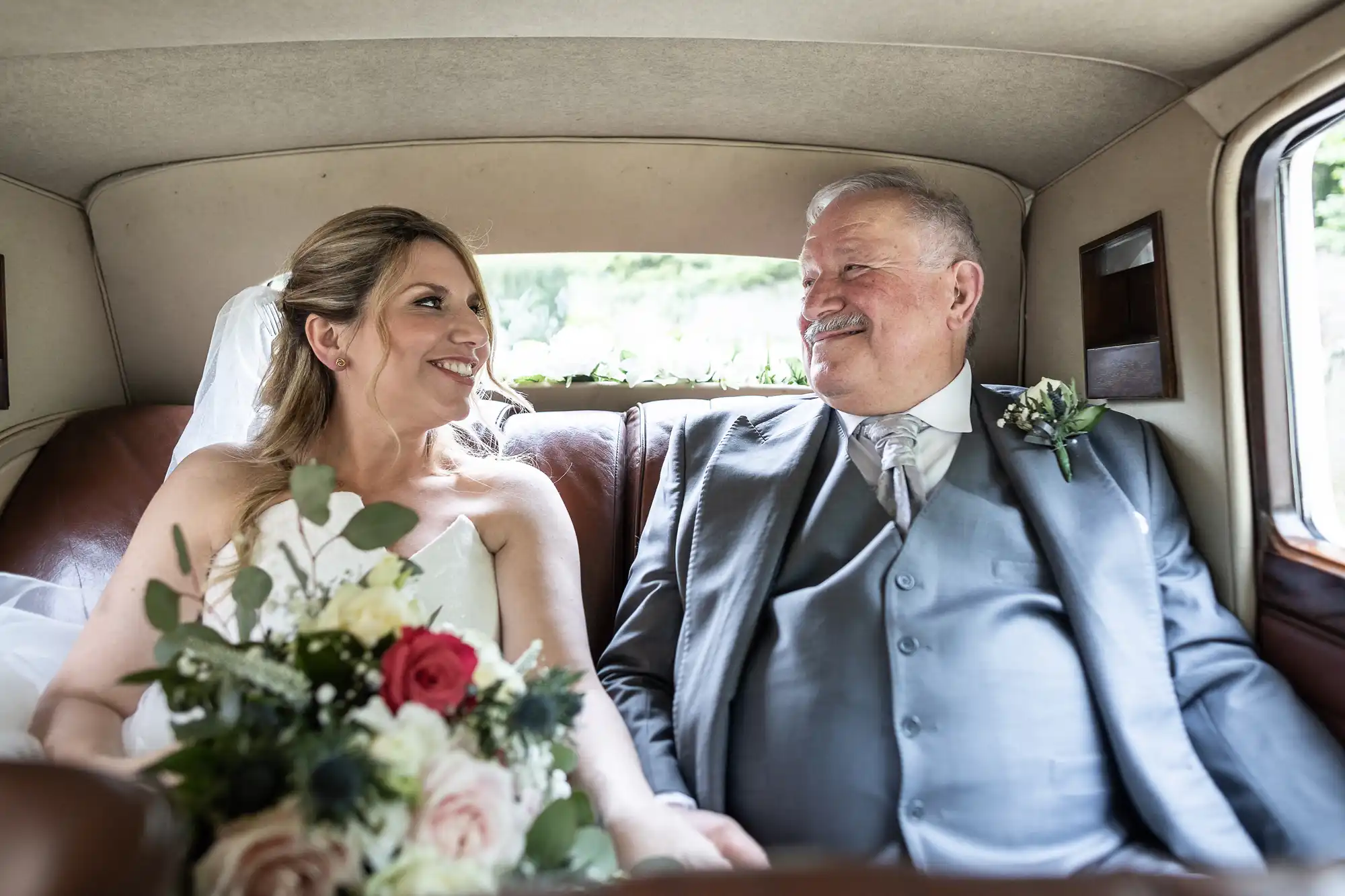 A bride and an older man are sitting in the back of a vintage car, smiling at each other. Both are dressed formally, and the bride is holding a bouquet of flowers.