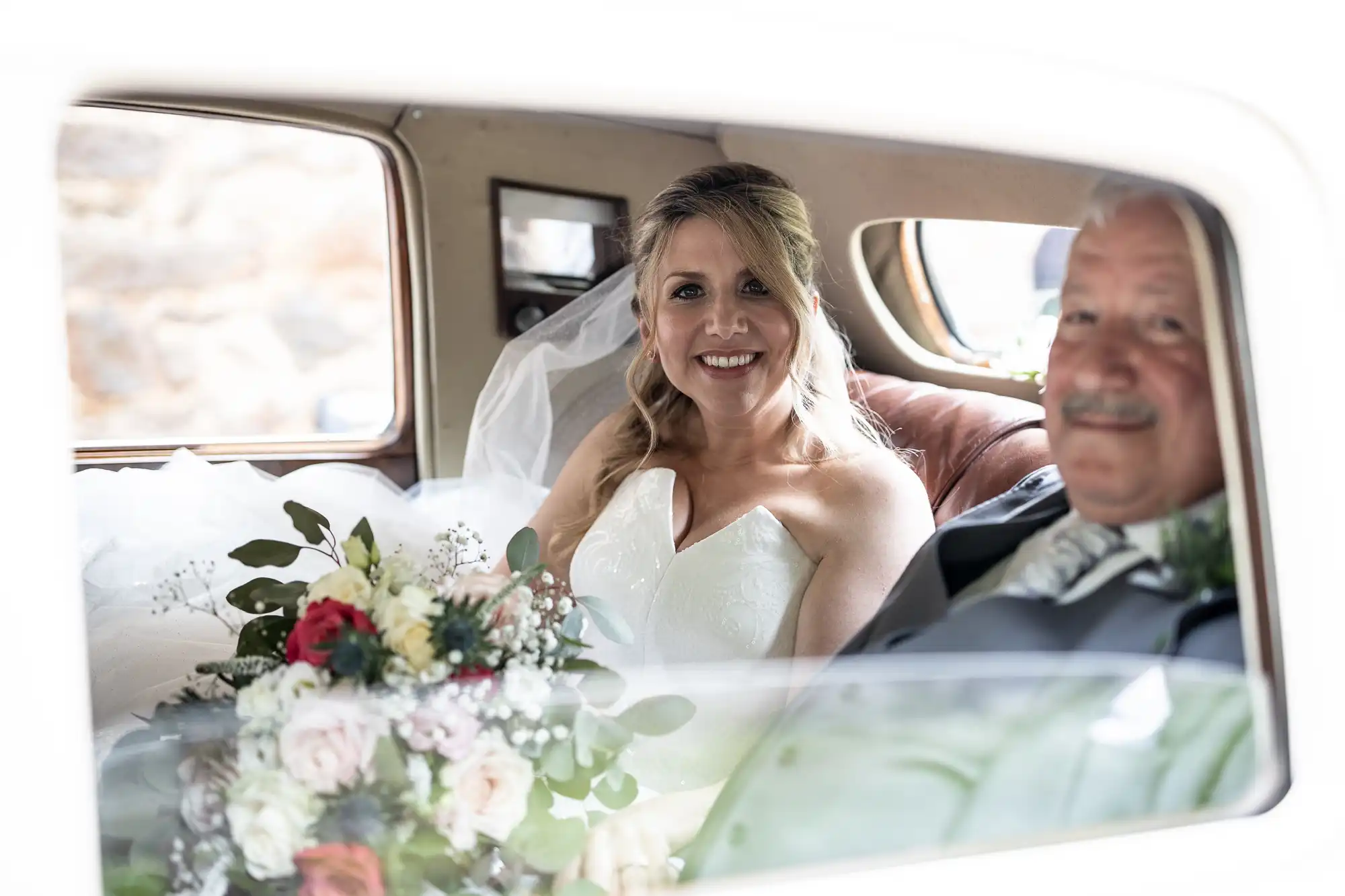 A bride in a white dress and an older gentleman in a suit sit inside a car. The bride holds a bouquet of flowers and smiles at the camera.