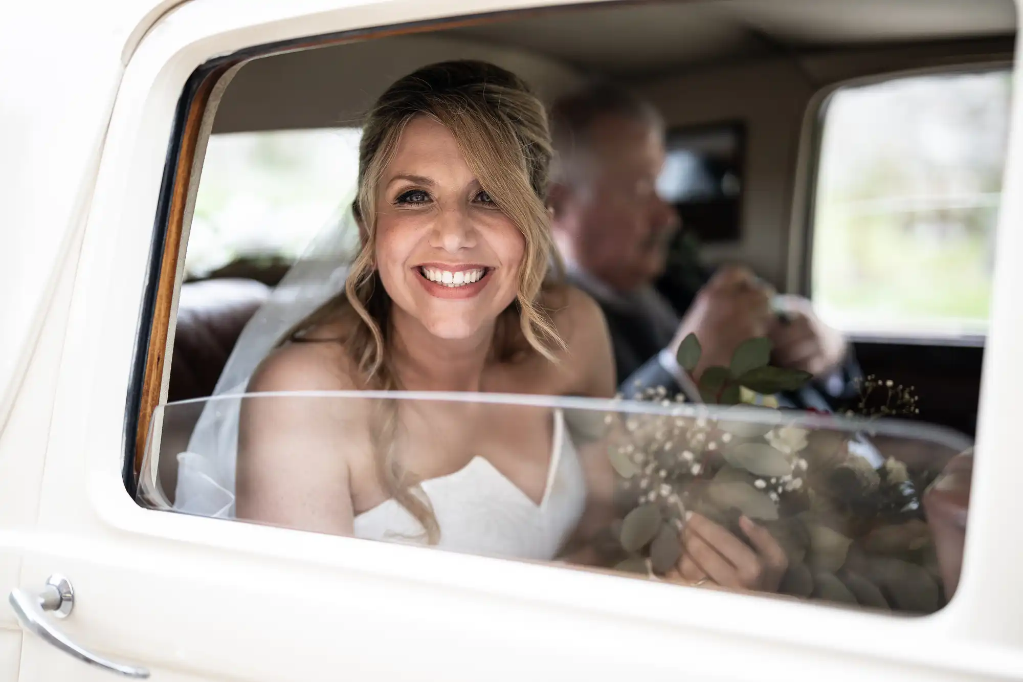 A smiling bride with blonde hair sits in the back of a car holding a bouquet, with an older man beside her.