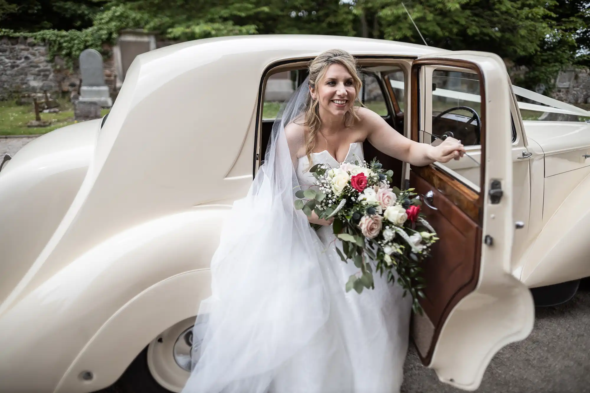 Bride holding a bouquet of flowers, stepping out of a vintage cream-colored car with the door open.