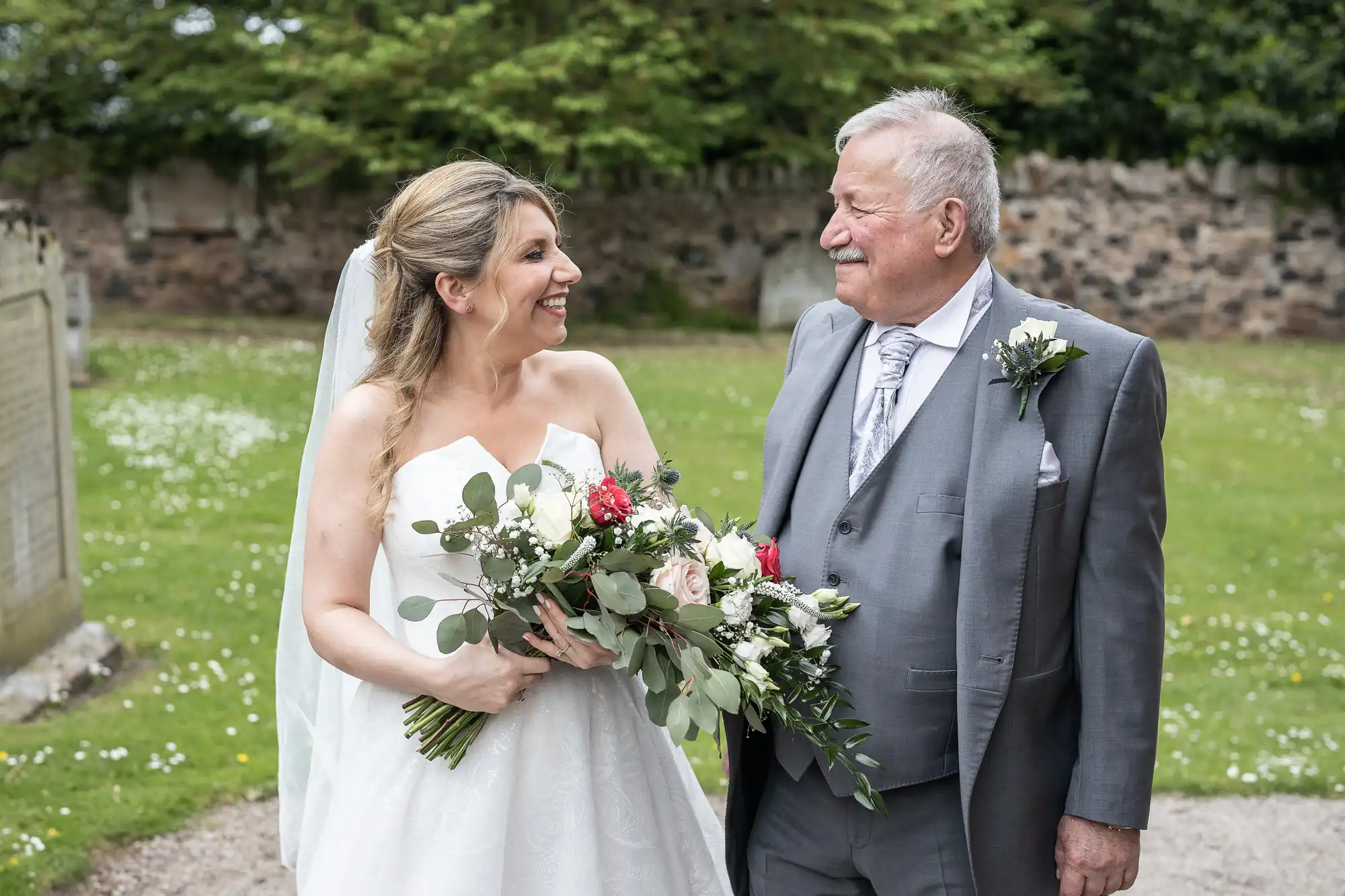 A bride in a white dress holds a bouquet and smiles at an older man in a grey suit with a boutonniere. They stand outdoors in front of green foliage and a stone wall.