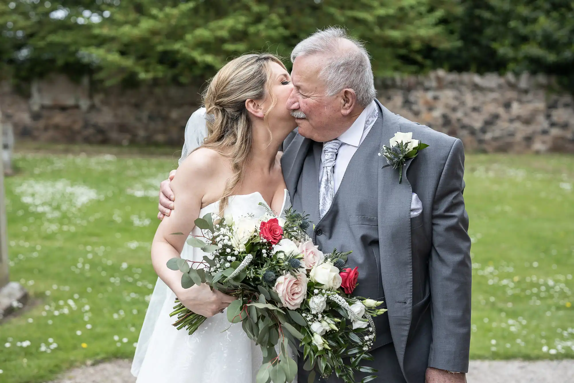 A bride in a white dress hugs and kisses an elderly man in a gray suit and tie. The bride is holding a bouquet of mixed flowers. They are outdoors with greenery and a stone wall in the background.