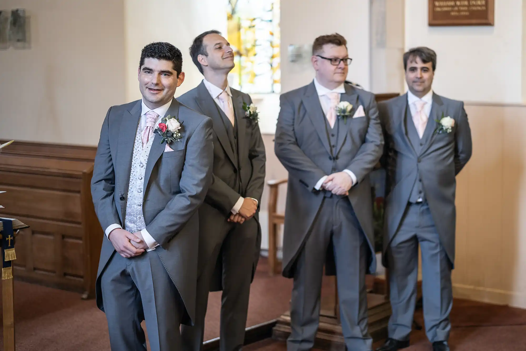 Four men in grey suits with boutonnieres stand together in a room with wooden paneling and a stained glass window. One man is smiling at the camera, while the others look in different directions.