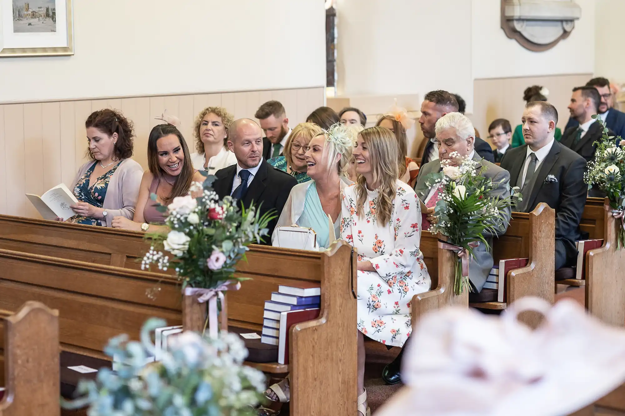 People are seated in pews inside a church, some are smiling and talking. Floral arrangements decorate the space, and the atmosphere appears light and cheerful.