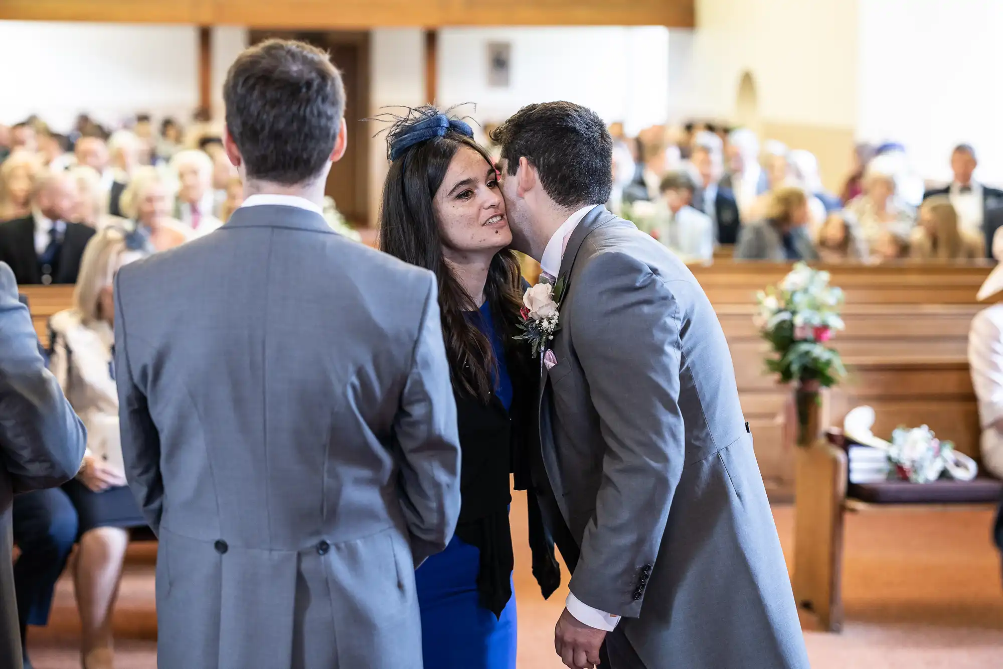 A man in a gray suit leans in to kiss a woman wearing a blue dress and a fascinator on the cheek in a crowded church. Another man in a gray suit stands facing them.