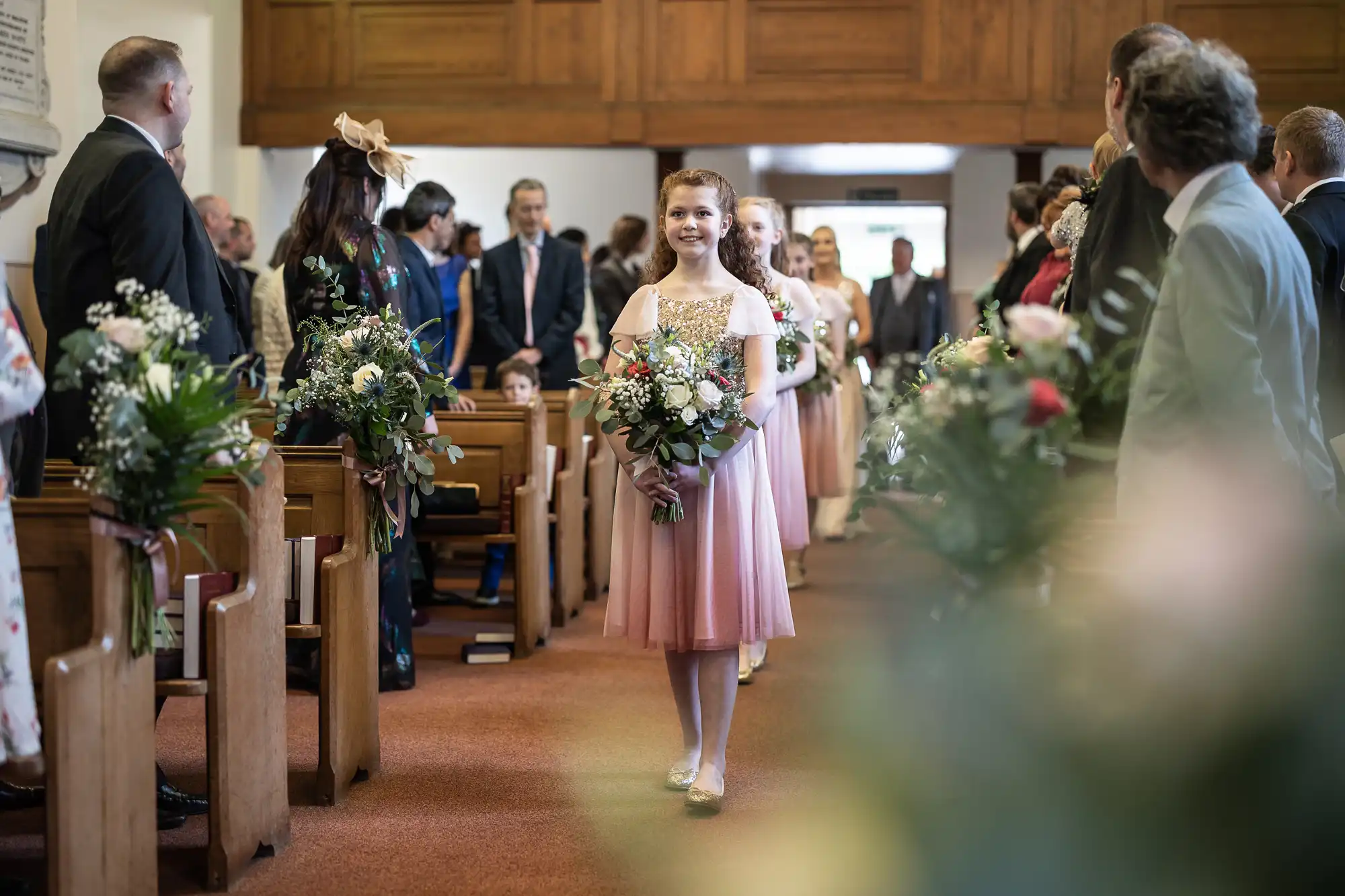 A young girl in a pink dress walks down the aisle carrying a bouquet of flowers at a wedding ceremony, surrounded by seated attendees.