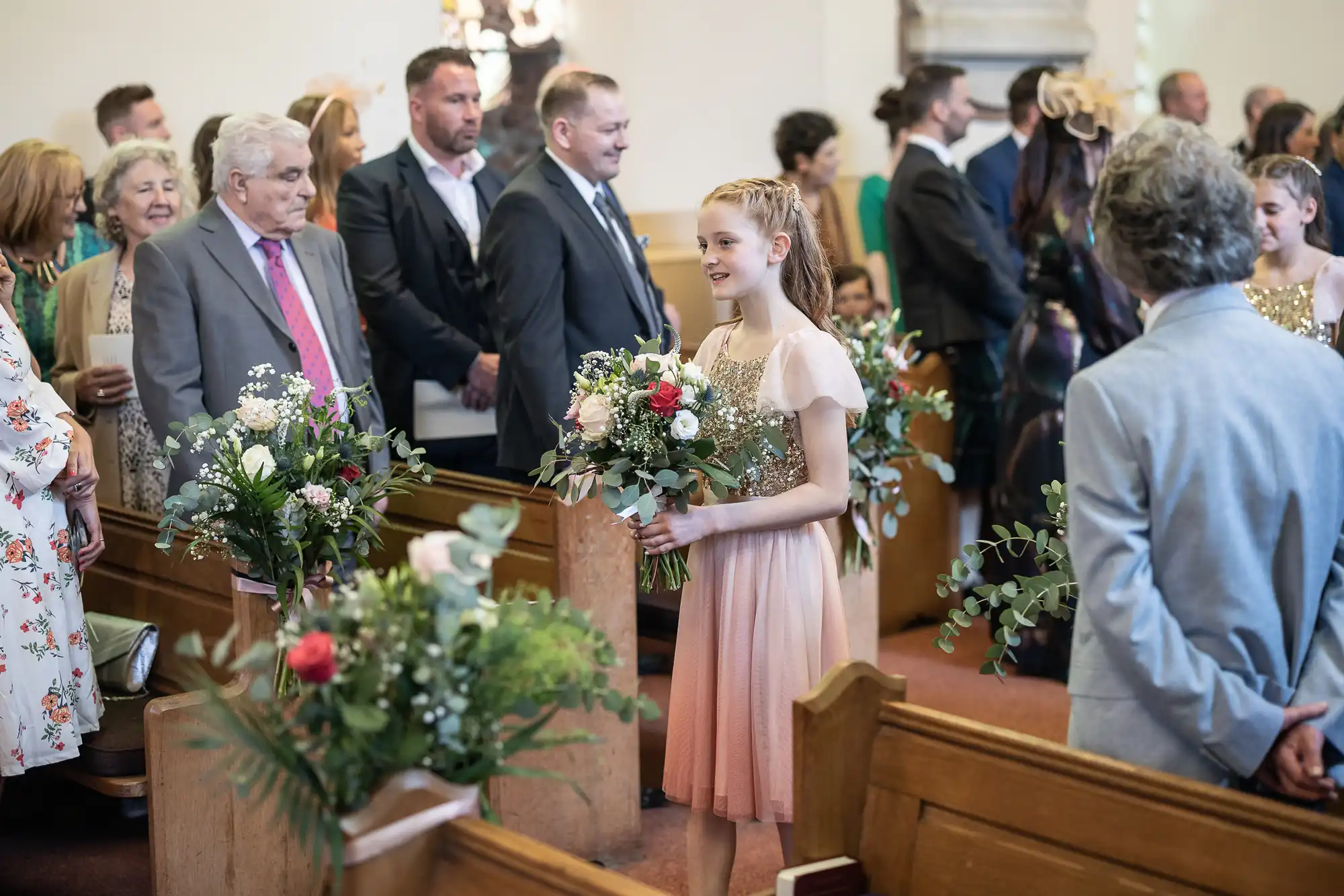 A young girl in a pink dress, holding a bouquet of flowers, stands in a church aisle. Guests in formal attire are seated and standing in rows of wooden pews, watching her.