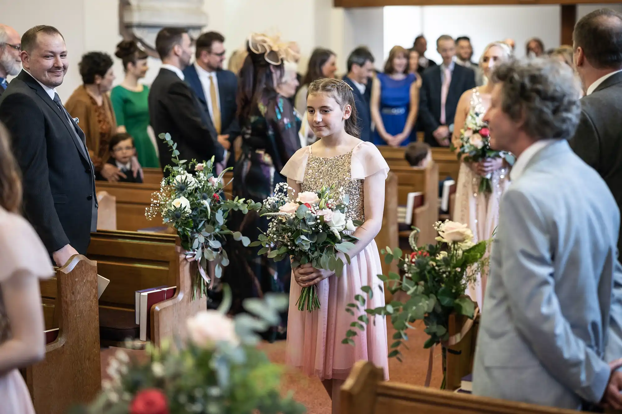 A girl in a pink dress holding a bouquet of flowers walks down the aisle of a church, surrounded by standing attendees in formal attire.