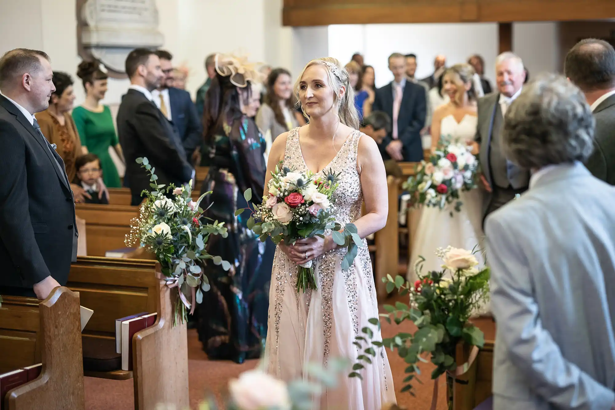 A woman dressed in a light gown holds a bouquet of flowers and walks down the aisle of a church, with guests seated and standing on either side, during a wedding ceremony.