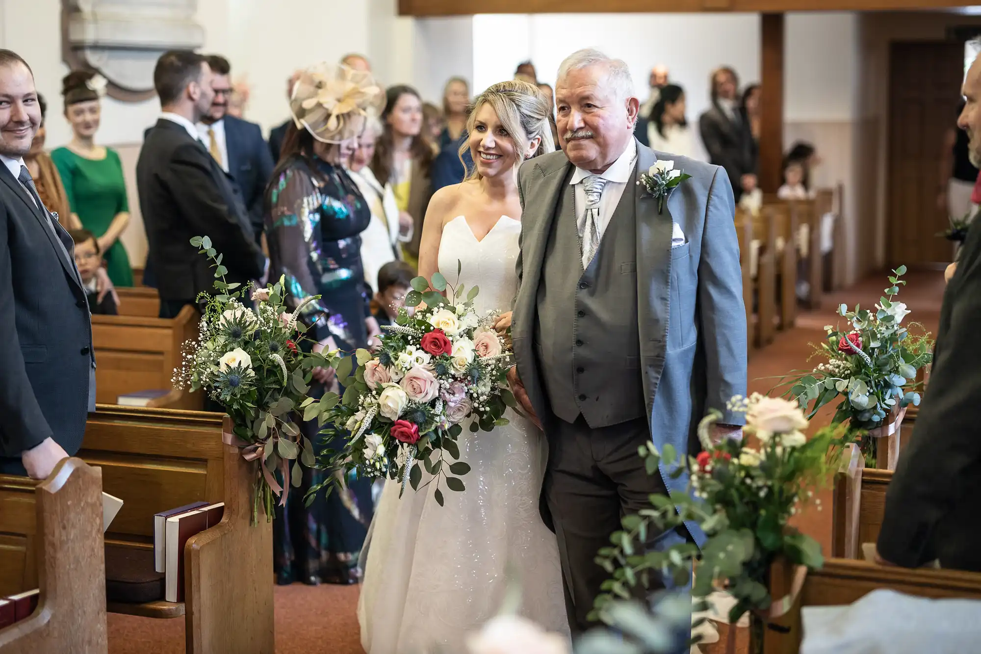 A bride in a white dress and a man in a gray suit walk down the aisle in a church, holding floral arrangements. People in formal attire are seated and standing, watching the procession.