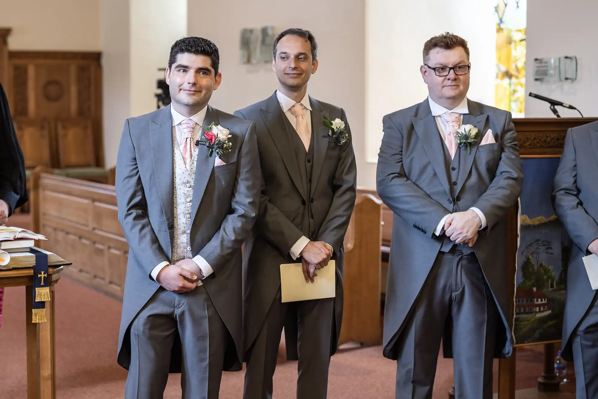 Three men in formal grey suits stand inside a church, smiling. Two hold papers; one stands with hands clasped.