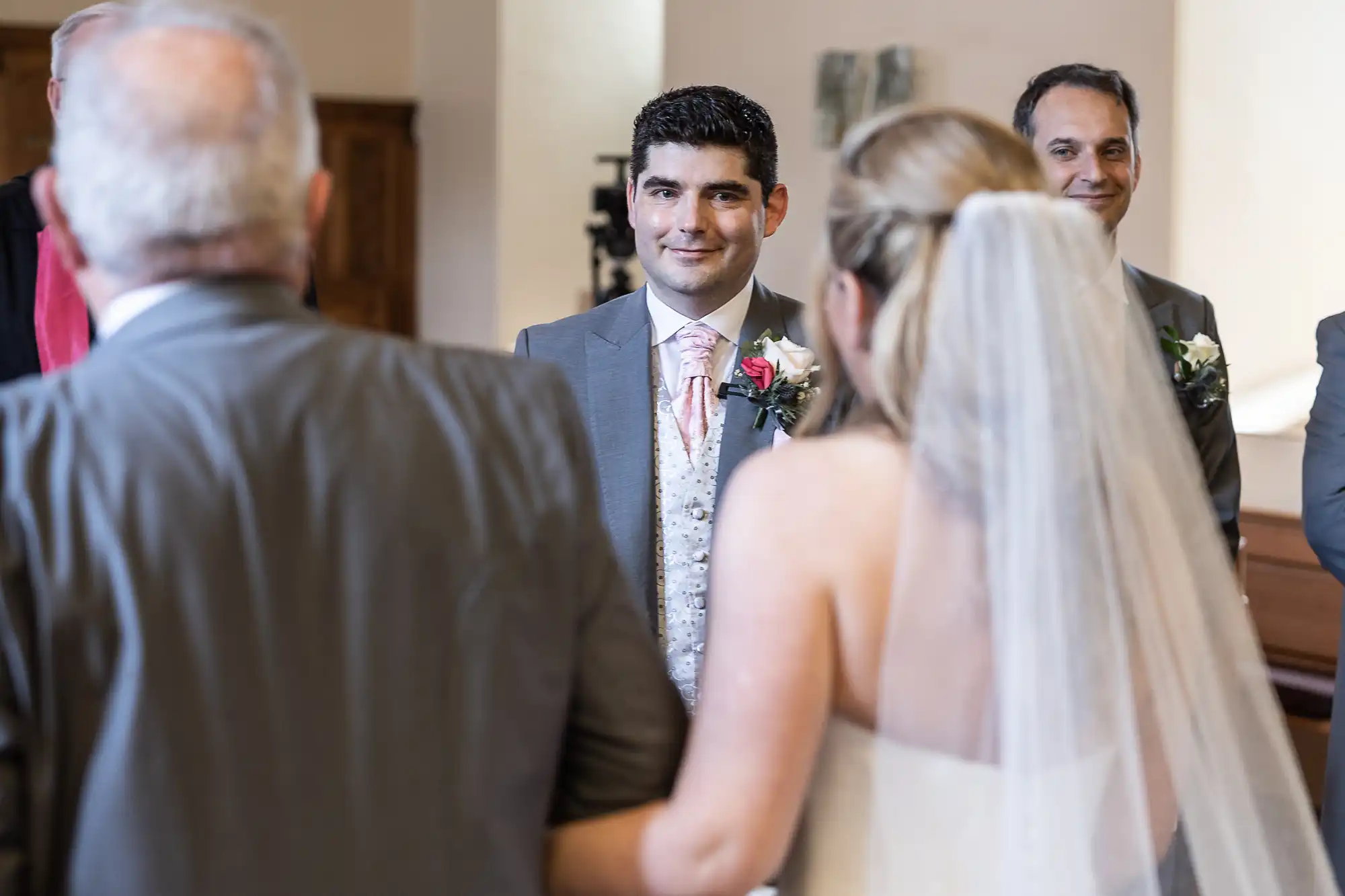 A bride and groom stand facing each other during a wedding ceremony. The bride holds onto an older man's arm and the groom smiles. Another man is visible in the background, also smiling.