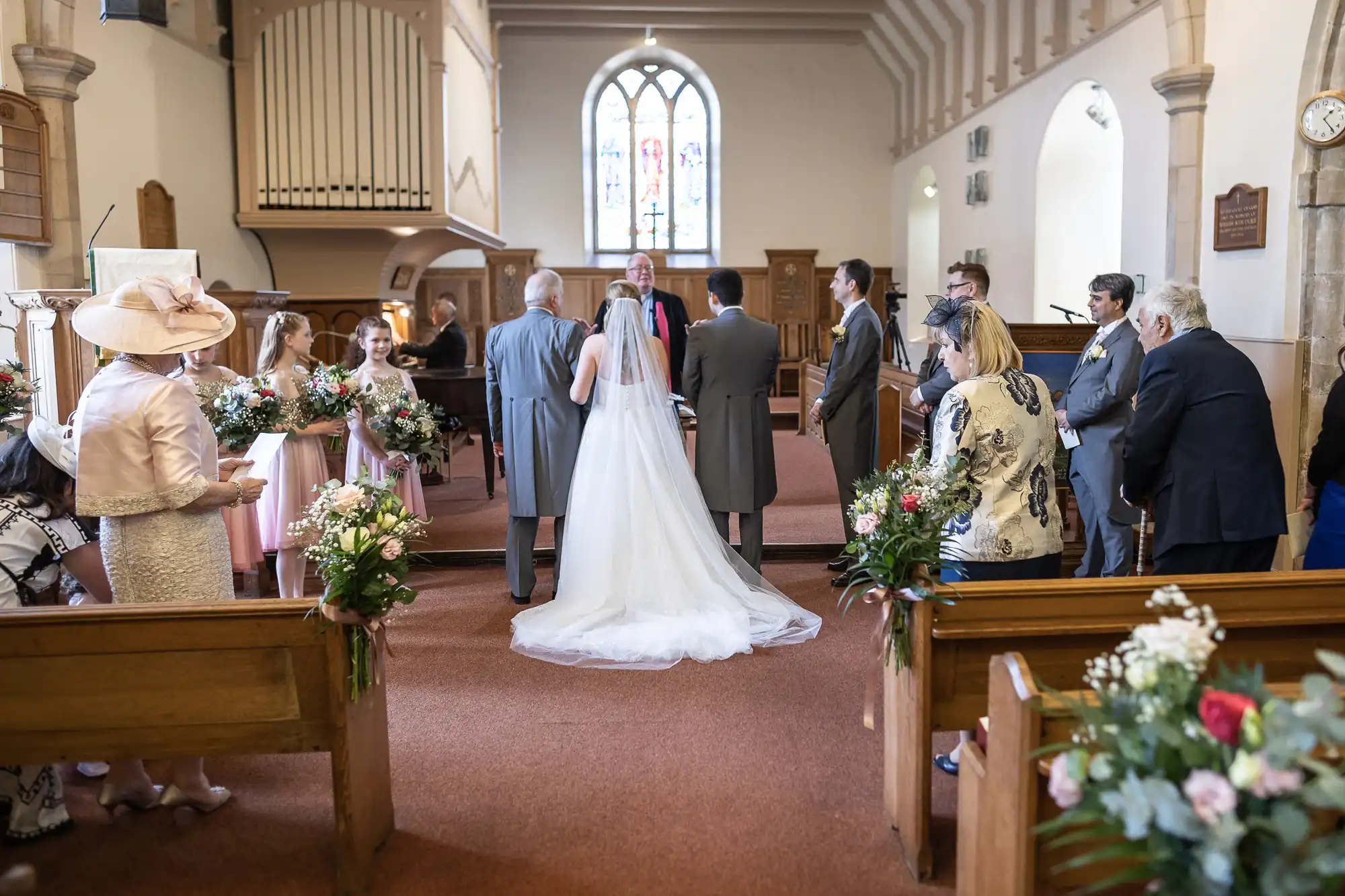 A bride and groom stand at the altar during a wedding ceremony in a church. Guests are seated and some stand around, dressed formally. Flower arrangements decorate the pews.