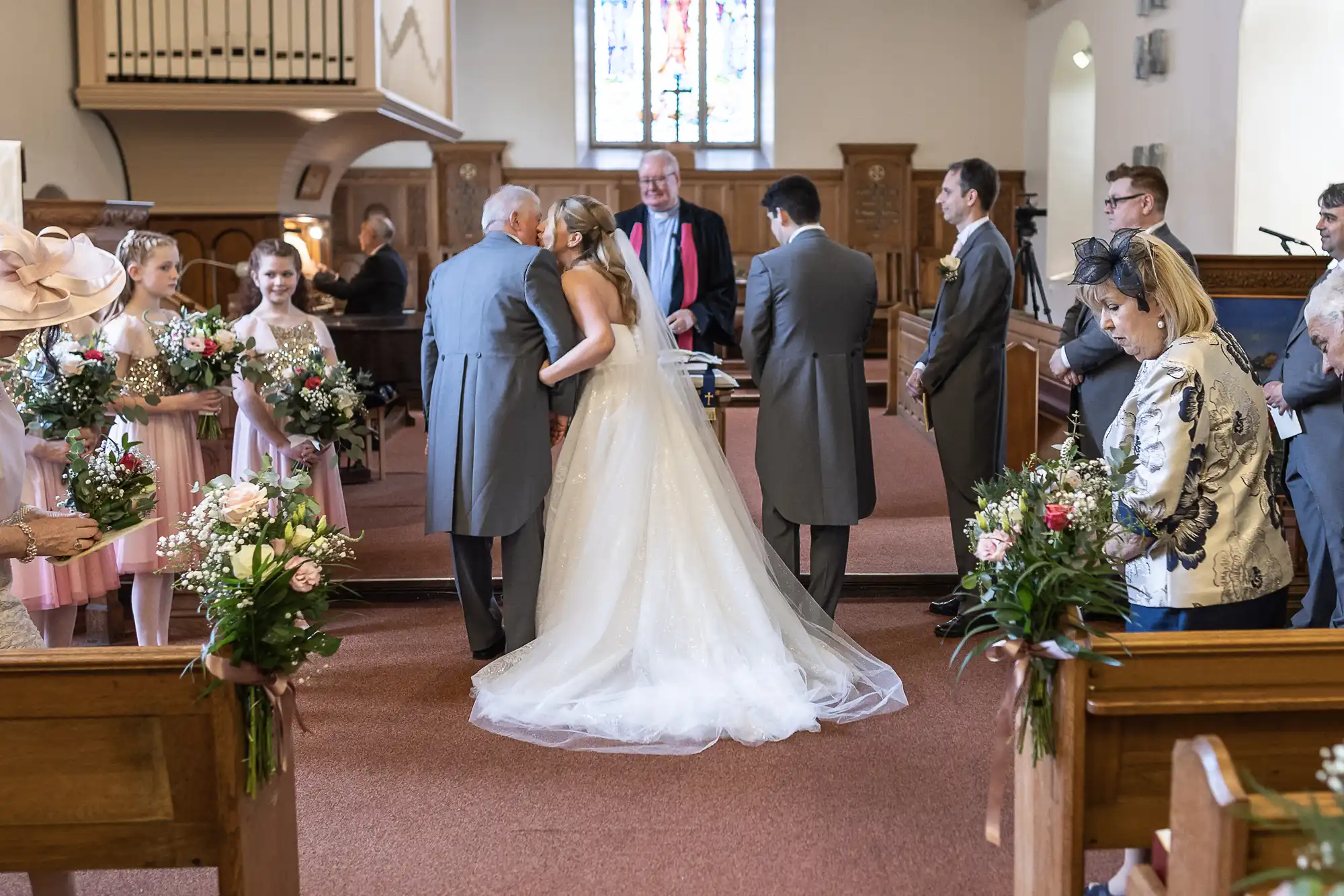 A bride in a white gown kisses an older gentleman while surrounded by wedding party members in a church. Guests and flower girls holding bouquets stand nearby.