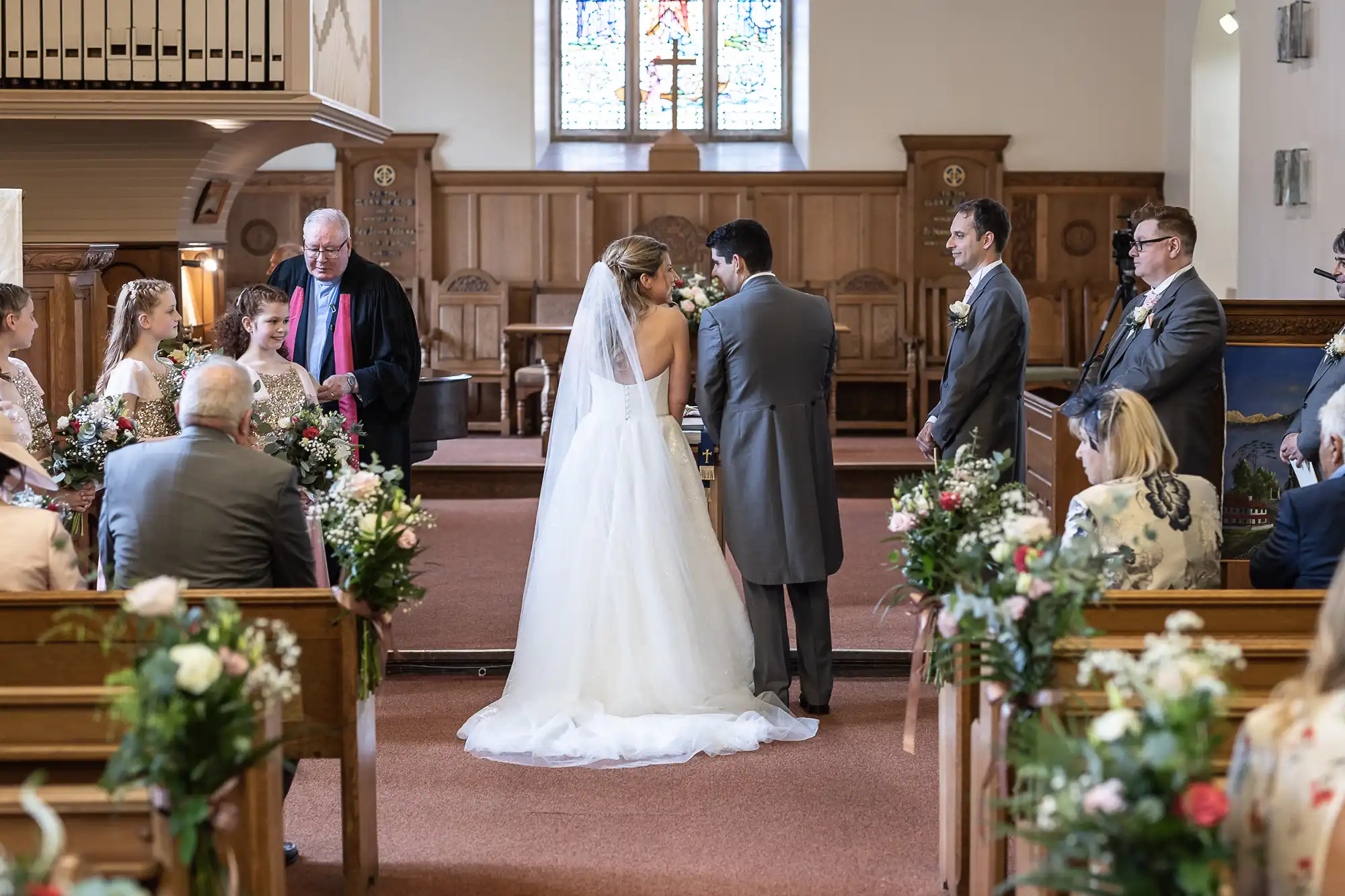 A bride and groom stand at the altar in a chapel, surrounded by their wedding party and guests. The officiant faces them as they hold hands, with floral arrangements lining the aisle.